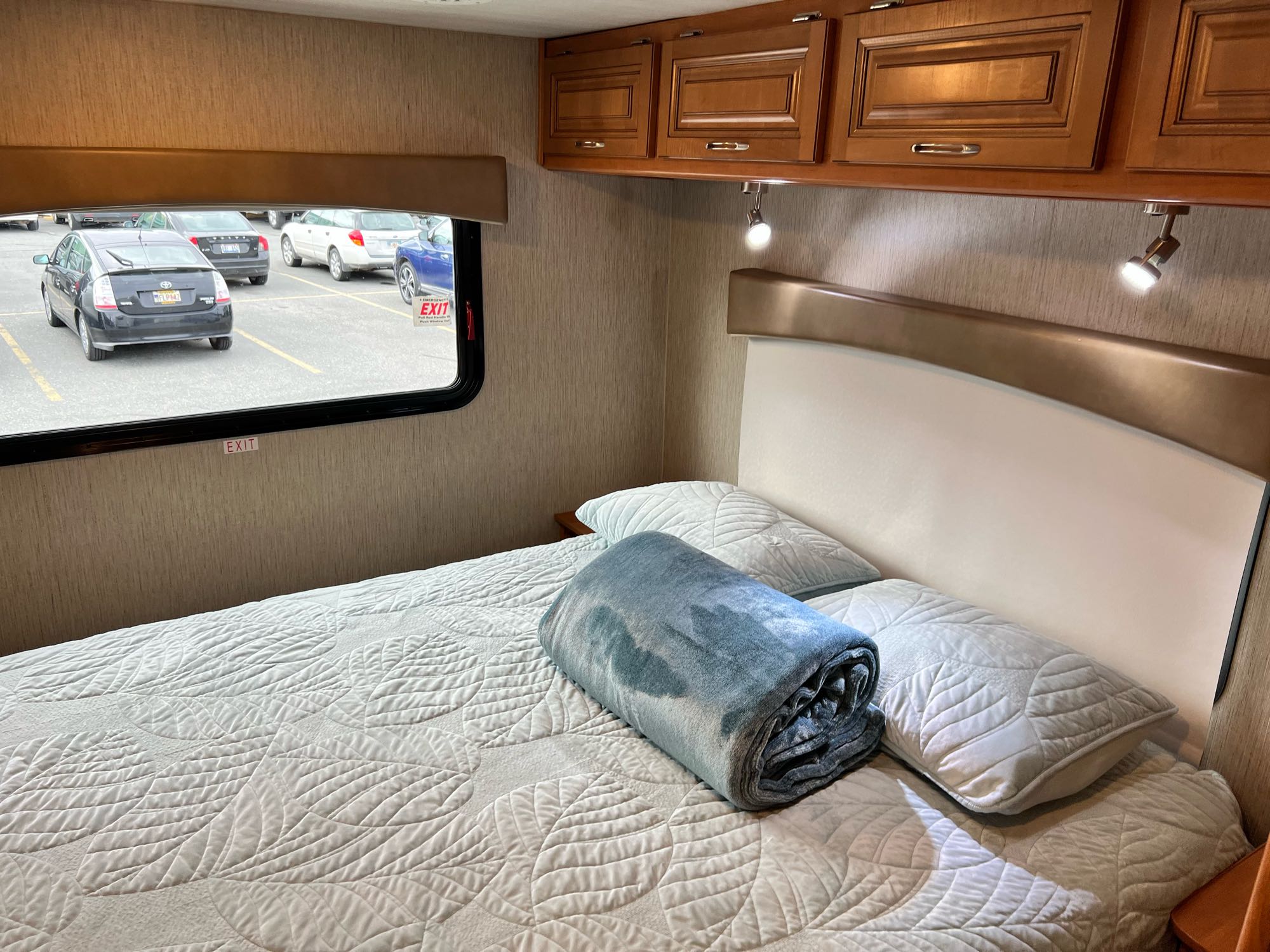 Cozy RV rental bedroom with quilted bedding, overhead storage, and a parking lot view from the window.. Nexus Phantom 32P 2019