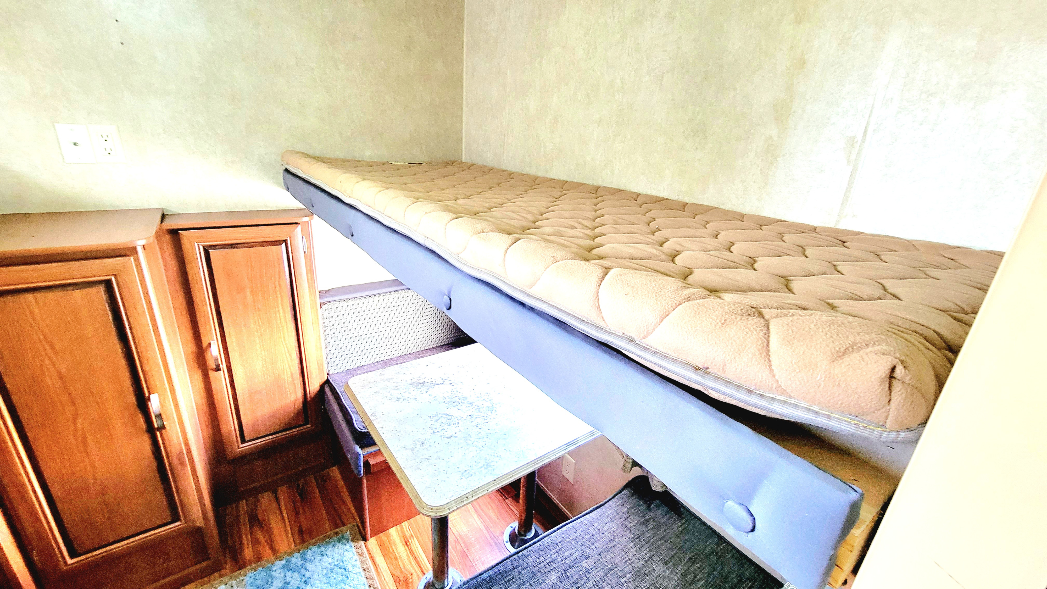 An RV rental interior showing a top bunk bed with a quilted mattress, a small table, and wooden cabinets.. Forest River Salem 2016