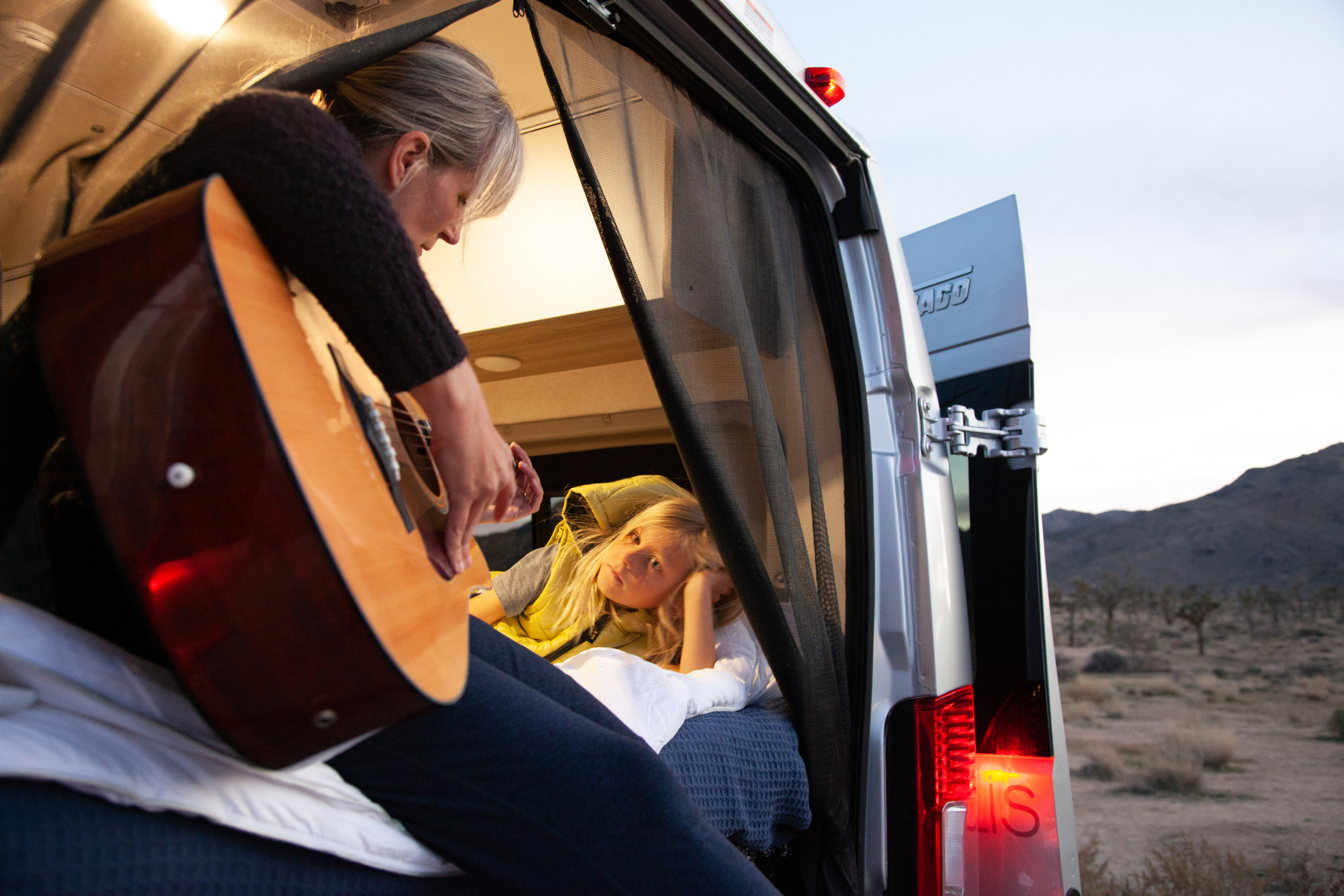 Woman plays guitar for a child inside an RV rental. The van's rear is open to a desert landscape.. Winnebago Solis 2023