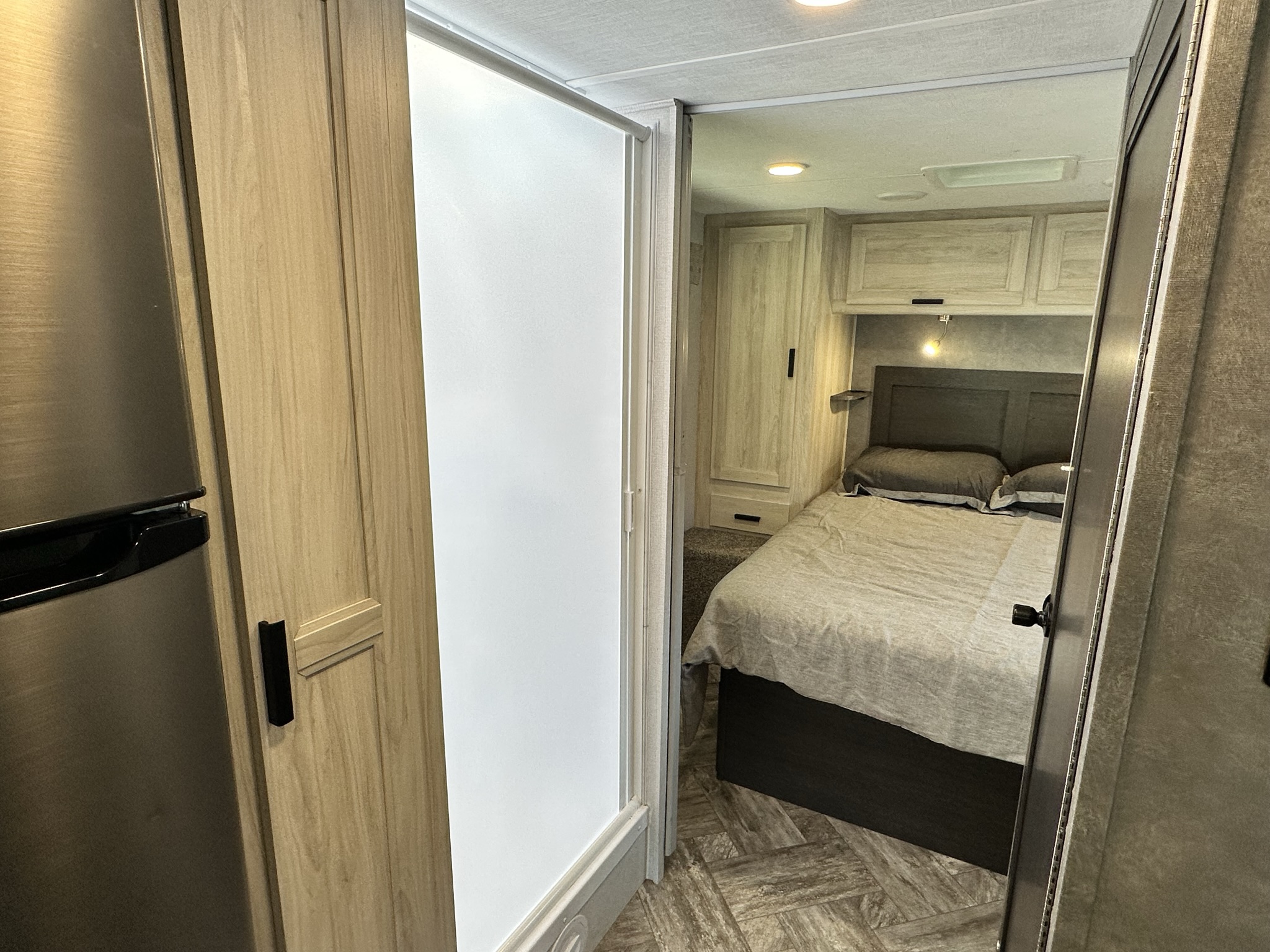 This RV rental interior showcases a cozy bedroom with overhead storage, a sleek shower door, and a modern refrigerator.. Coachmen Sunseeker 2022