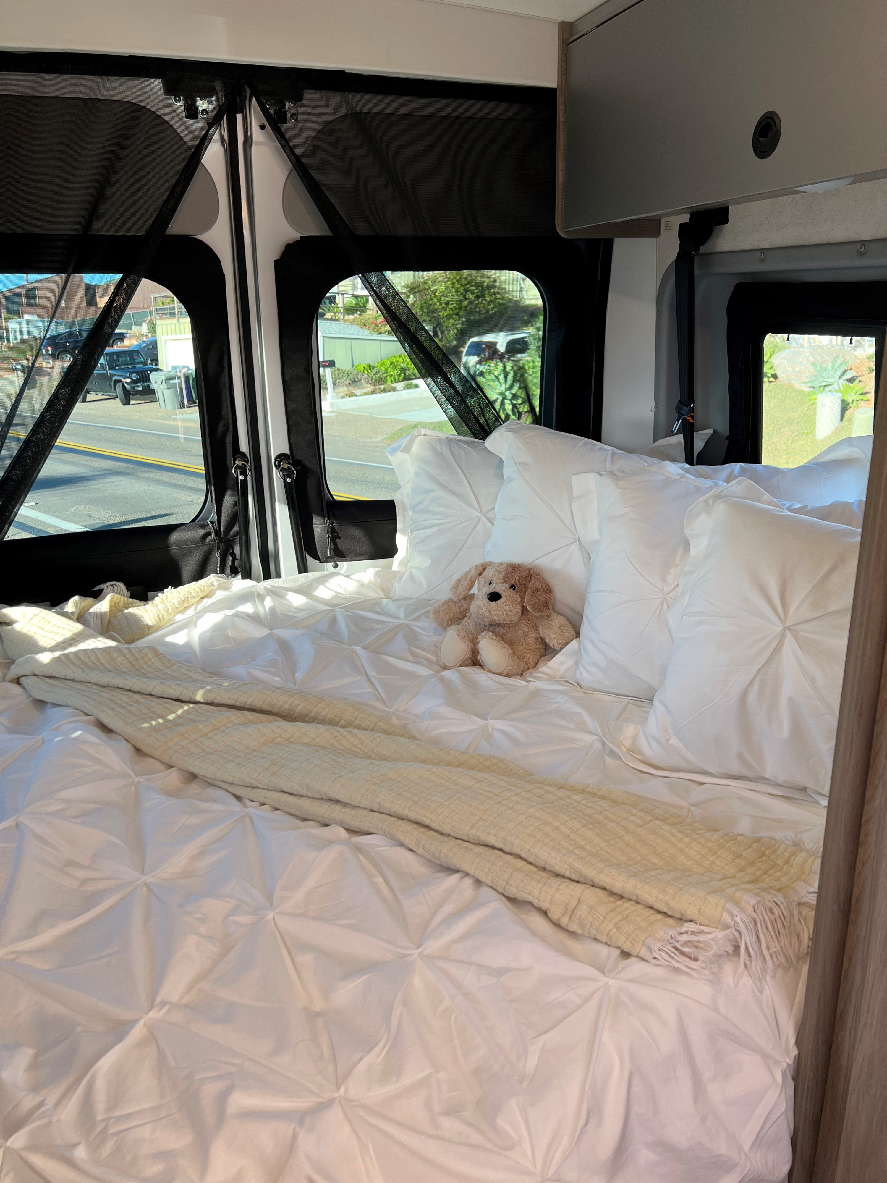 Inside an RV rental: a plush bed with white bedding, pillows, and a cute dog, overlooking an outdoor scene.. Winnebago Solis 2022