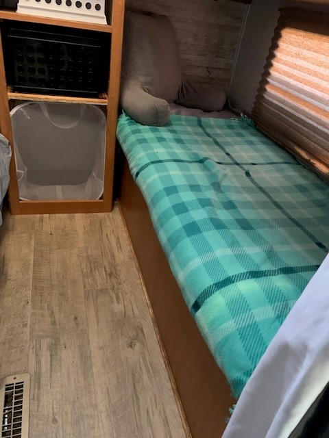 Cozy RV rental bedroom with plaid blanket, backrest pillow, shelving holding a crate & laundry hamper.. Pilgrim International Inc Pilgrim 2007