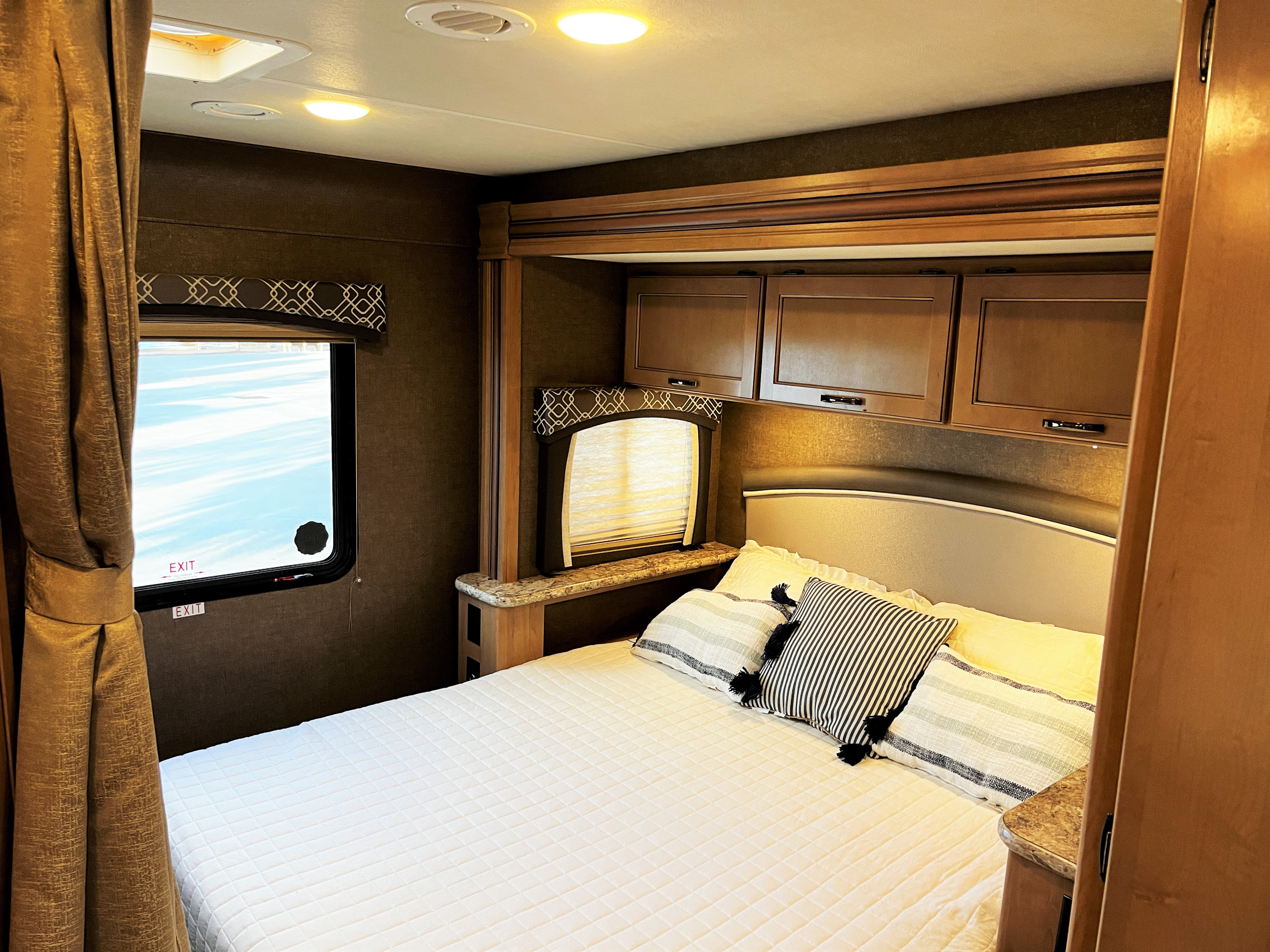 Comfortable RV rental bedroom features a quilted bed, overhead cabinets, and window with curtains. Ready for adventure!. Thor Chateau 2019