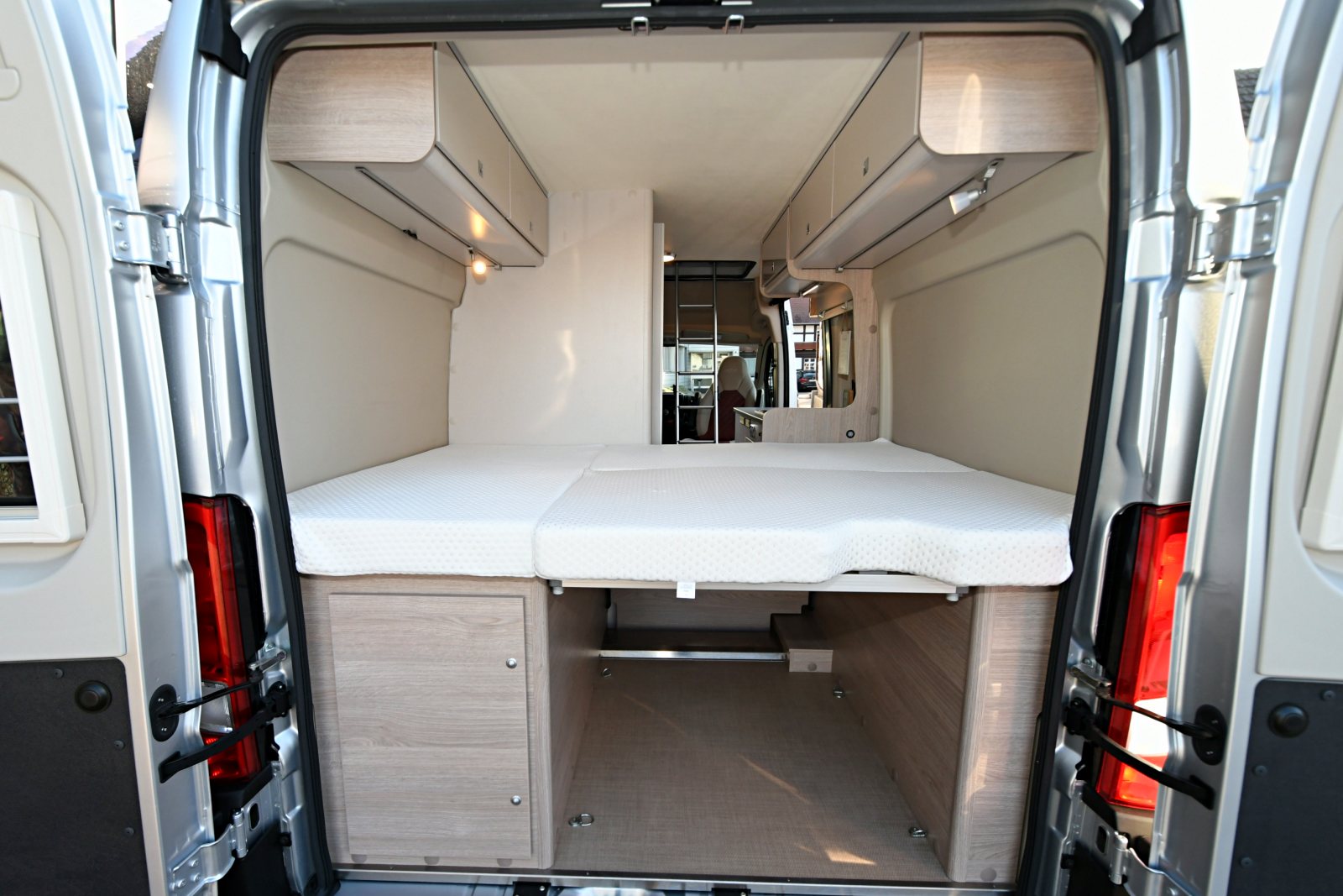 Spacious RV rental interior: elevated rear bed with under-bed storage and overhead cabinets, viewed from the open back doors.. Bürstner Campeo 600 2023