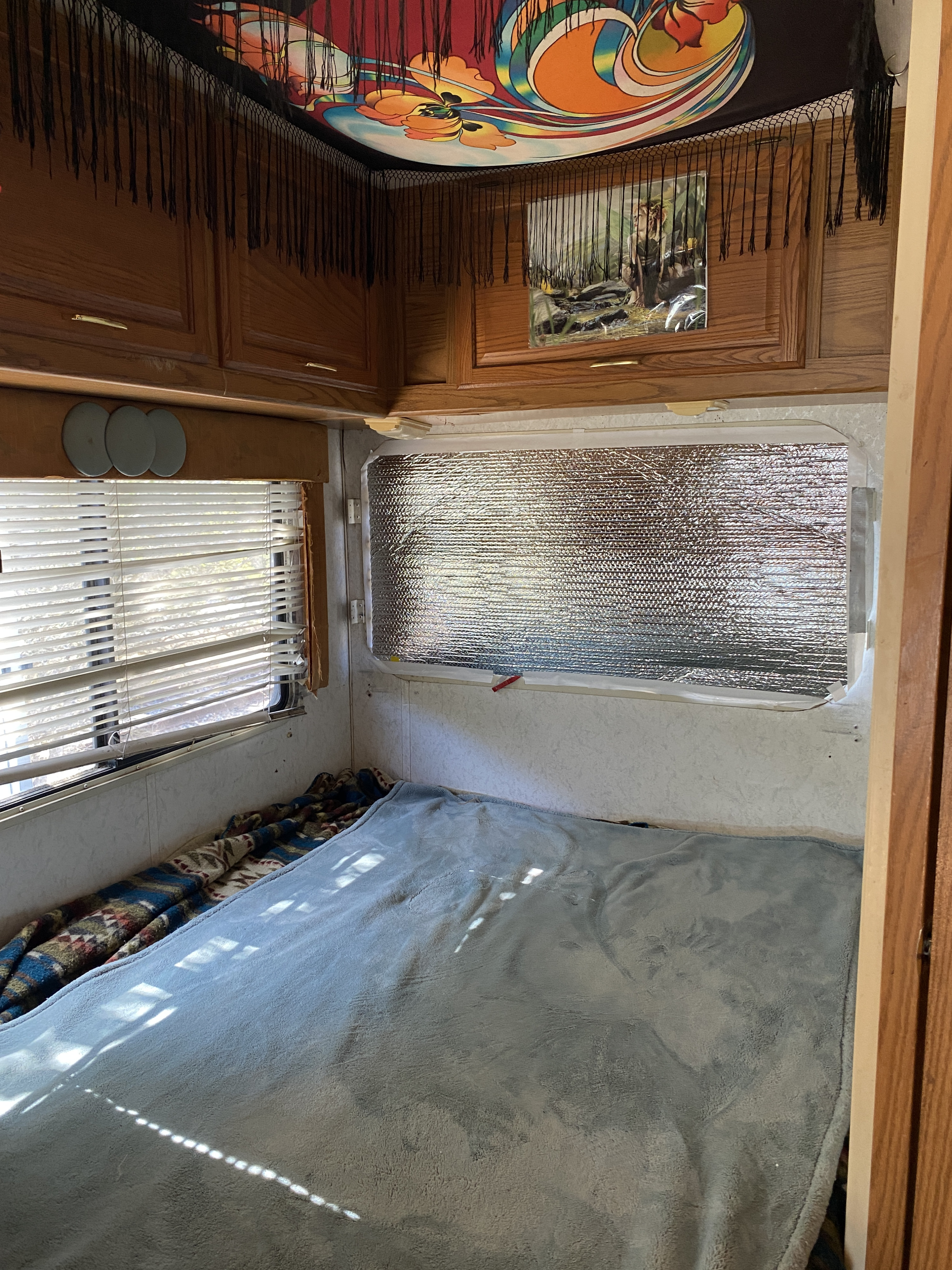 Bright RV rental bedroom: bed, overhead wood cabinets, blinds, reflective window insulation, and colorful tapestry.. Winnebago Chalet 2000