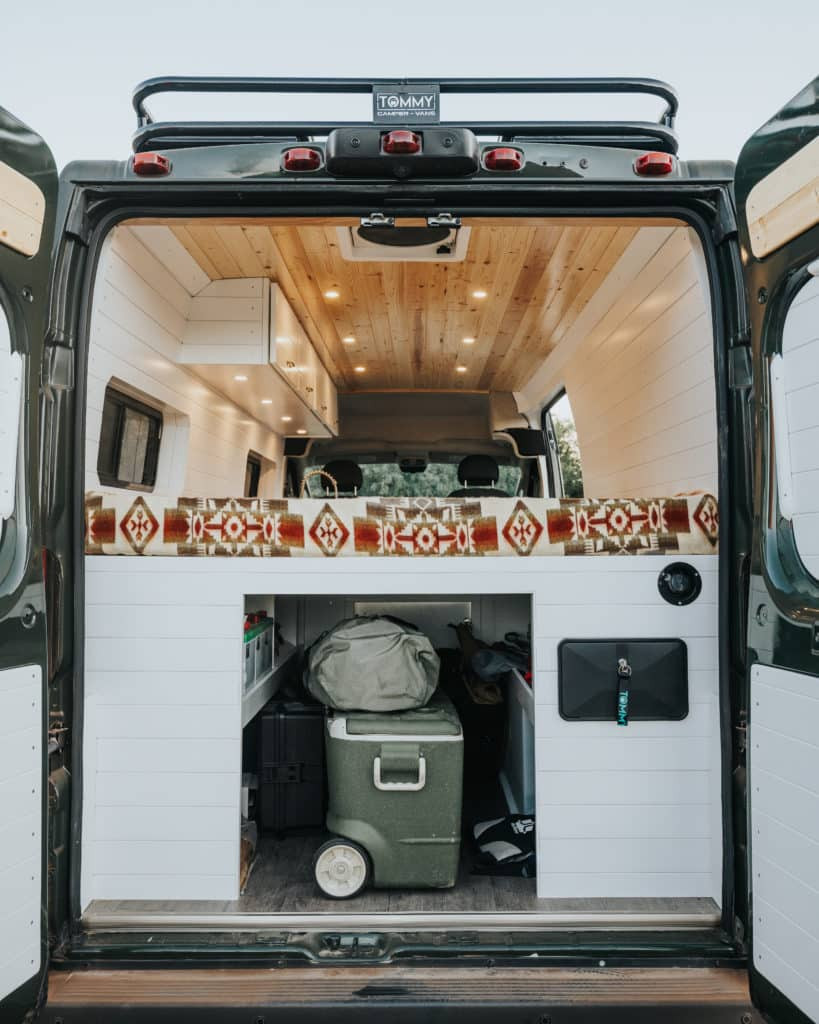 Experience comfort in this custom RV rental's spacious interior: bed with patterned blanket, storage area with cooler, and wooden finishes.. Dodge Sprinter 2018