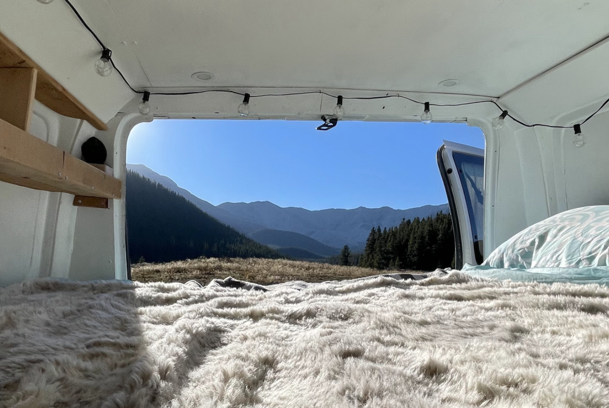 Cozy rv rental interior: A fluffy bed, string lights, and open door reveal sunny mountains and a forest view.. Ford Econoline 2012