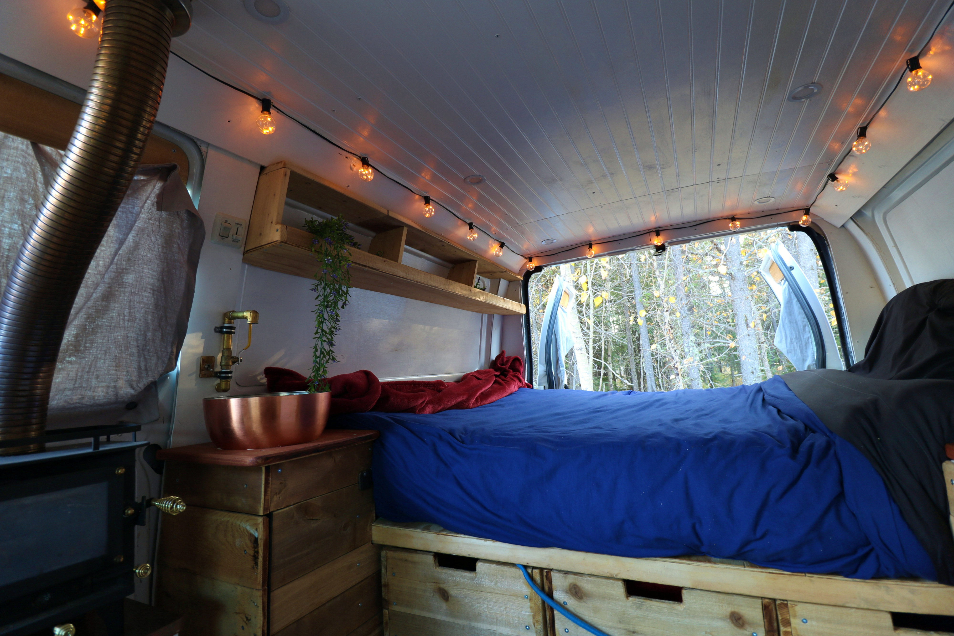 Cozy RV rental interior: bed, wood stove, copper sink, and string lights, offering a serene forest view for van life.. Ford Econoline 2012