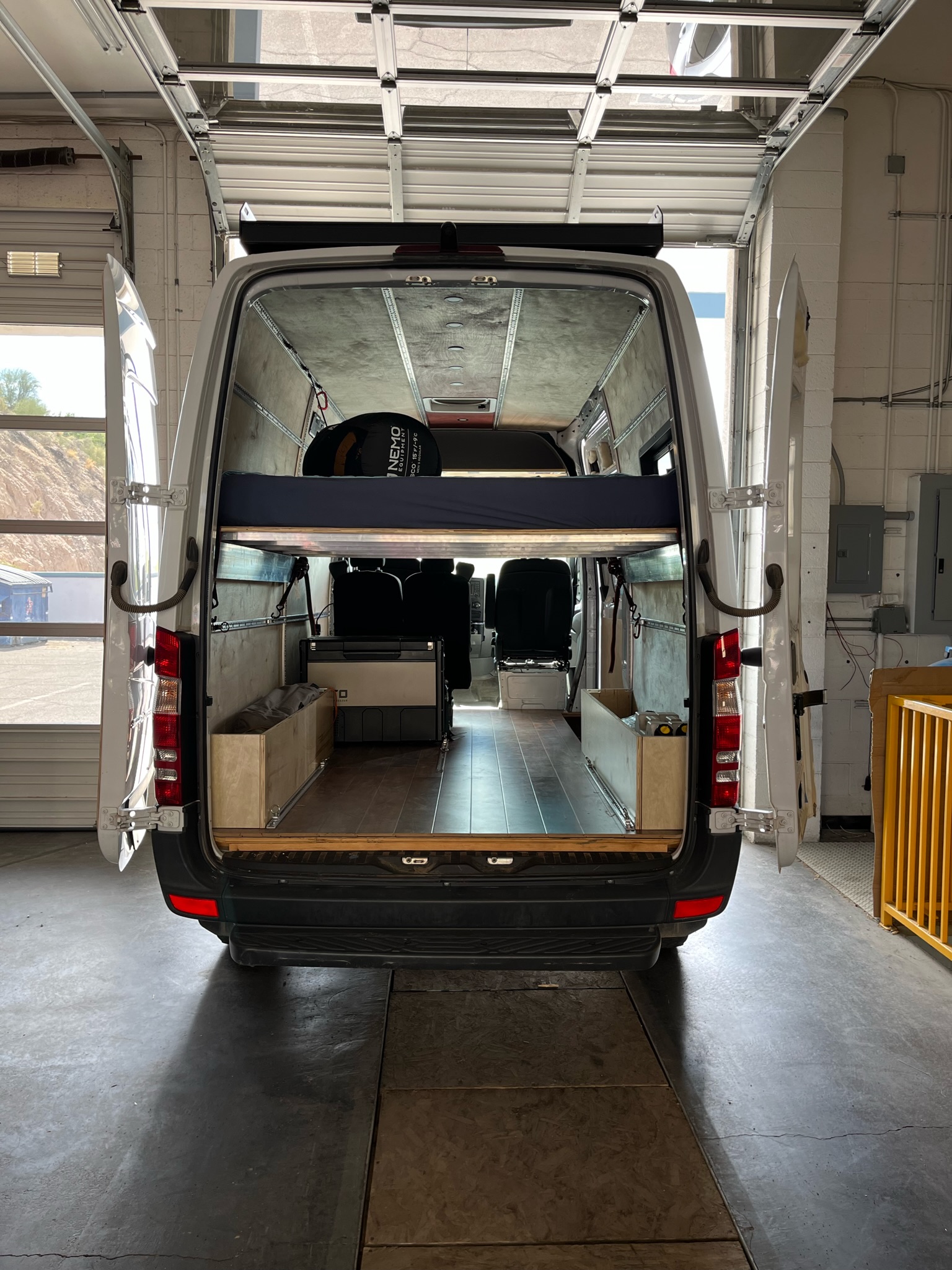 Interior of an RV rental camper van, showing a bed, fridge, and storage, parked in a garage with open doors.. Mercedes-Benz Sprinter 2017