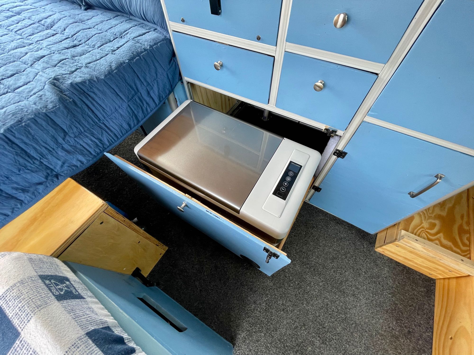 RV rental interior: Pull-out refrigerator visible under custom blue cabinets next to a comfy bed.. Dodge Ram Promaster 1500 2021