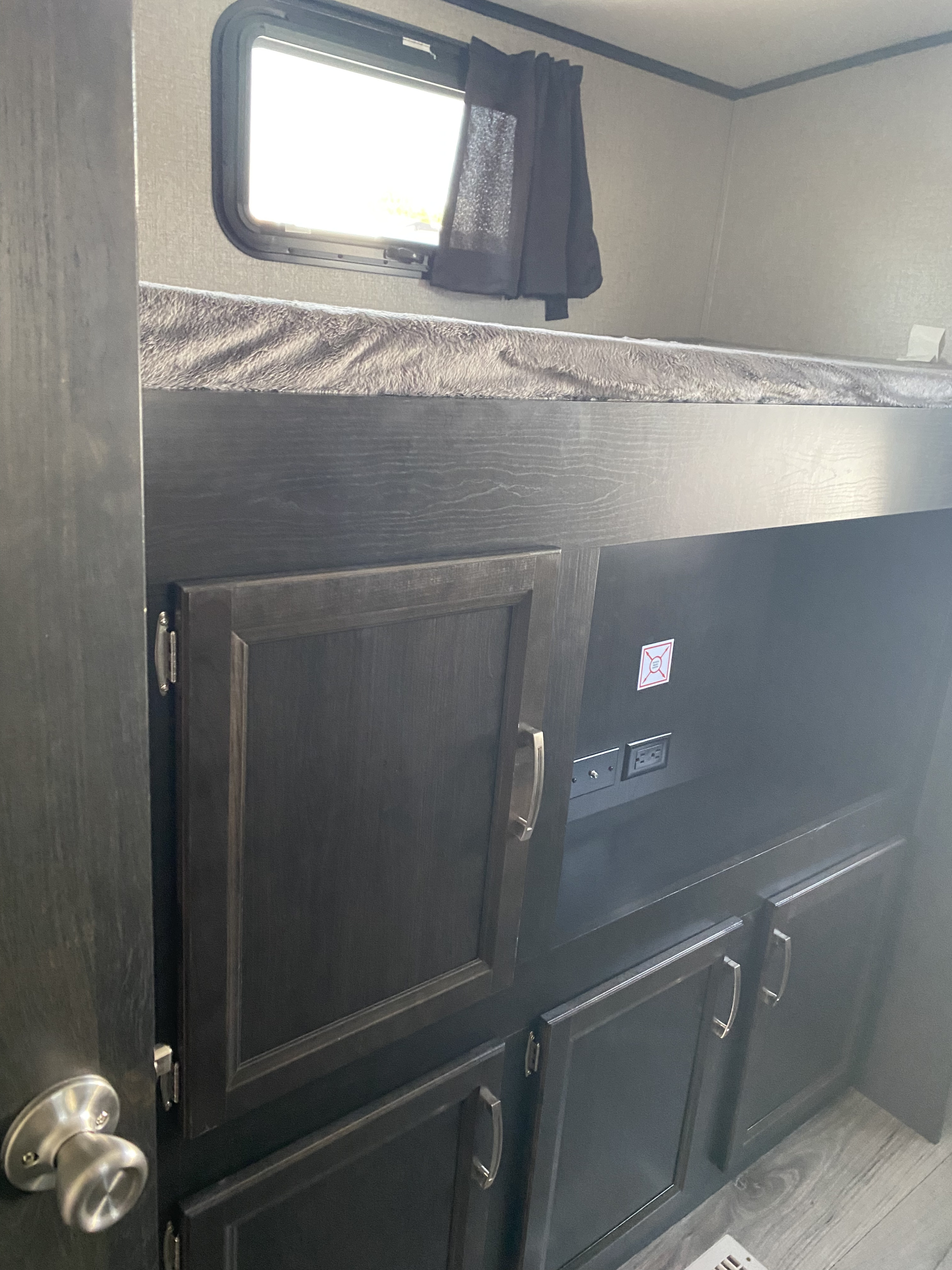 View inside an RV rental showing a bunk bed, dark wood cabinets with storage & outlets, and a window with a curtain.. Jayco White Hawk 2021