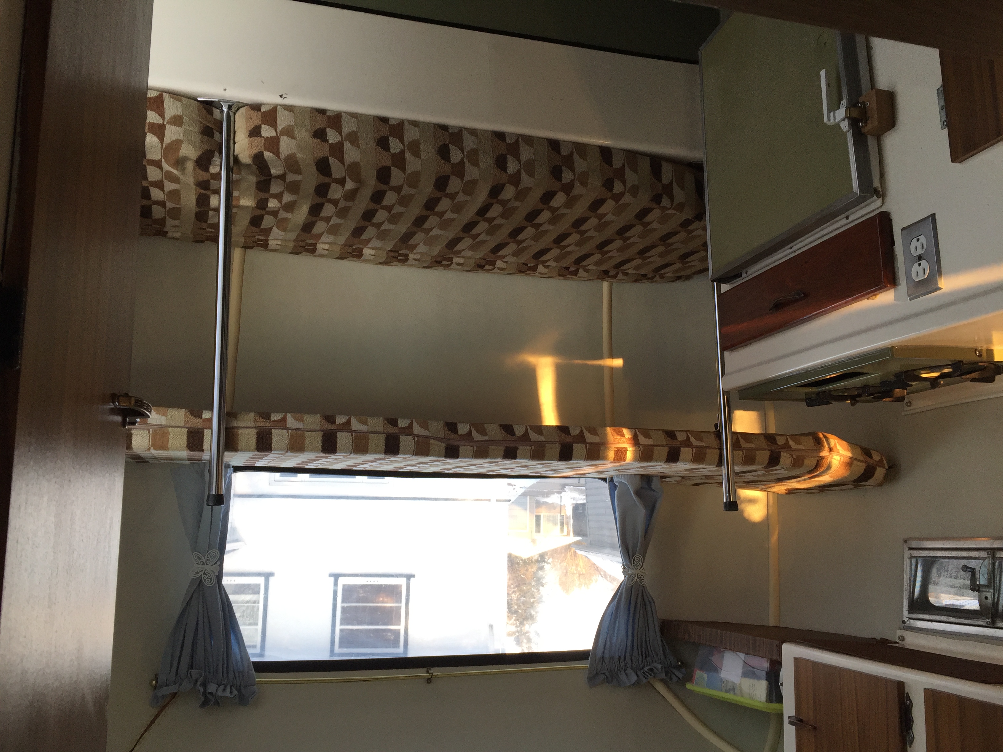 Interior view of an RV rental: two bunk beds with patterned cushions, a window with curtains, and a visible kitchen section.. Boler B13 1973