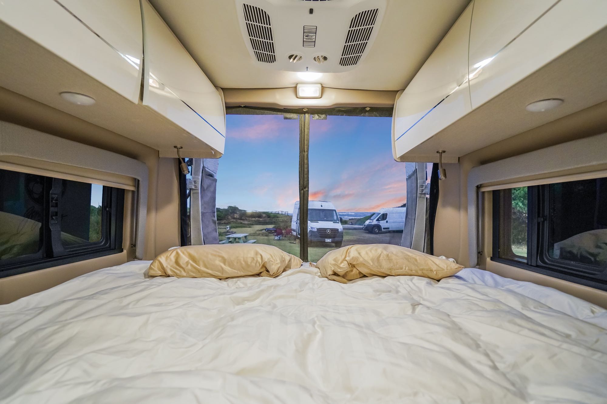 Experience ultimate comfort in this cozy RV rental bedroom, featuring a large bed and a scenic view of a campsite and sunset.. Thor Tellaro 2023