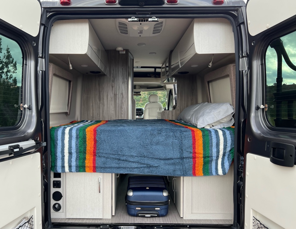 Inside this RV rental: a neatly made bed with a striped blanket, overhead storage, and under-bed space for luggage.. Thor Motor Coach Tellaro 2022