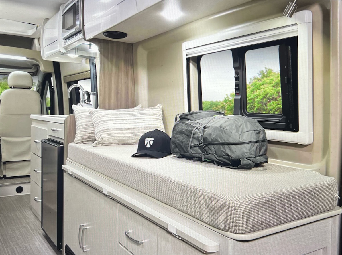 Step inside this modern RV rental! A comfortable bench with pillows and travel gear, a compact kitchen, and a scenic window offer a relaxing space.. Thor Motor Coach Tellaro 2022