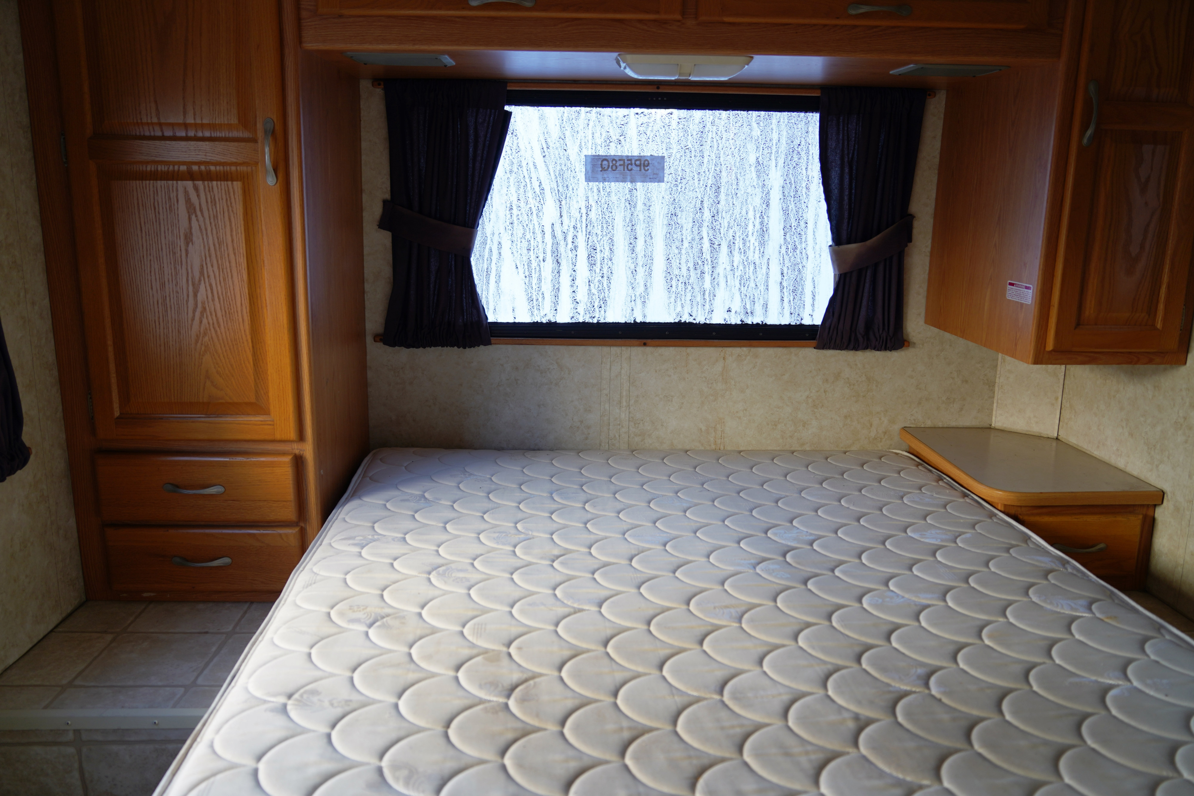 Cozy RV rental bedroom: white mattress, wood storage, window with rain streaking, and dark curtains.. Majestic 28A 2006