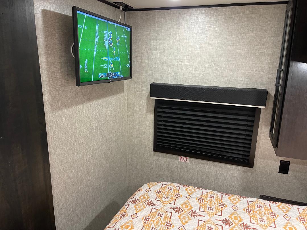 Inside an RV rental bedroom: a mounted TV displays a football game, above a window with blinds & a patterned bed.. Jayco Jay Flight 2022
