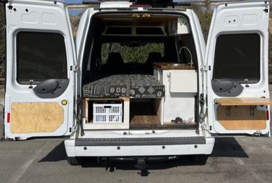 Rear of an open white camper van, showcasing a cozy bed, smart storage, and a sink setup. Ideal for an RV rental.. Ford Transit 2012