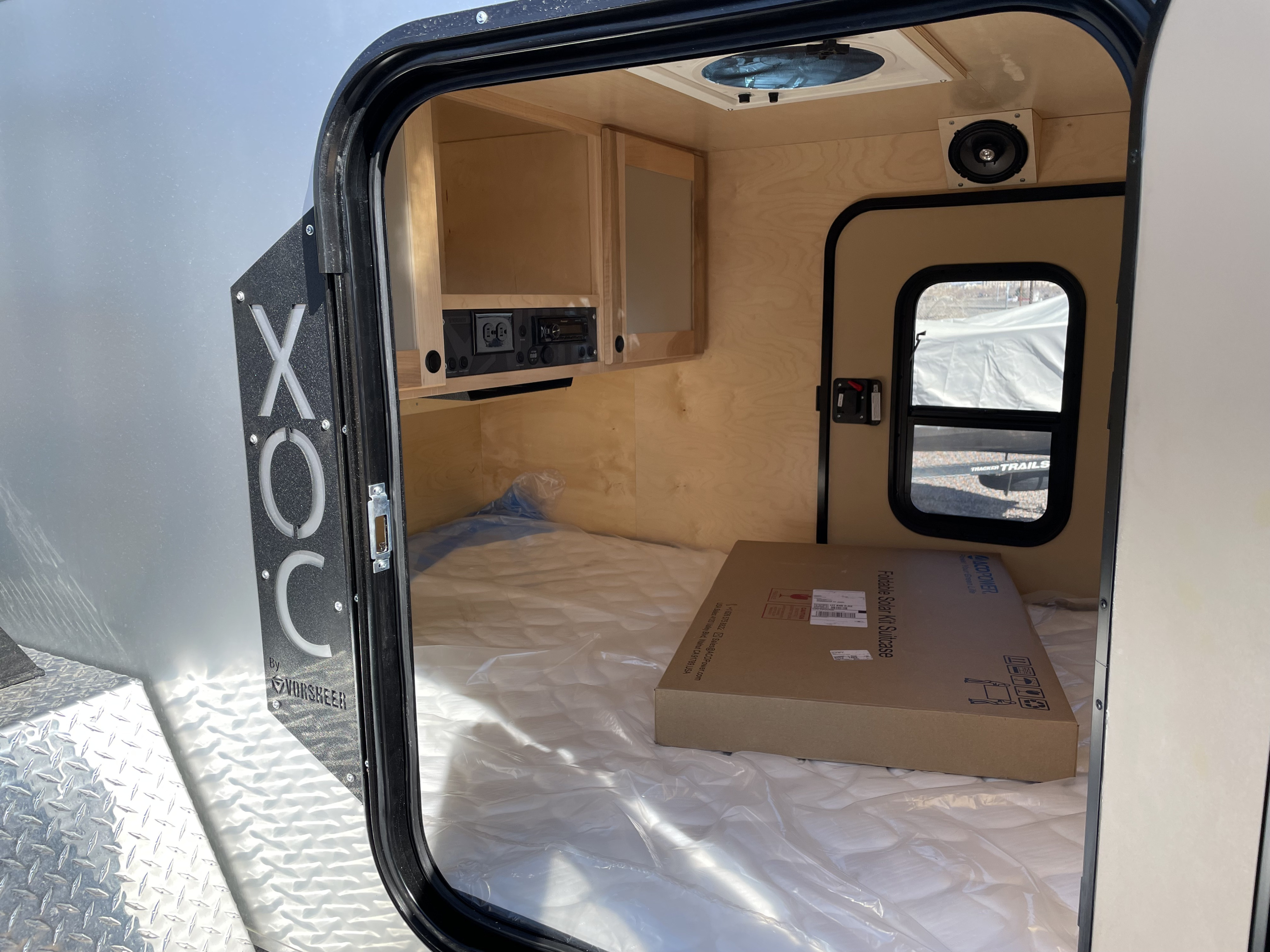 Interior of a compact rv rental camper, showing a mattress, wooden cabinetry, an overhead fan, and a new "Foldable Solar Kit Suitcase" box.. Vorsheer XOC 2022