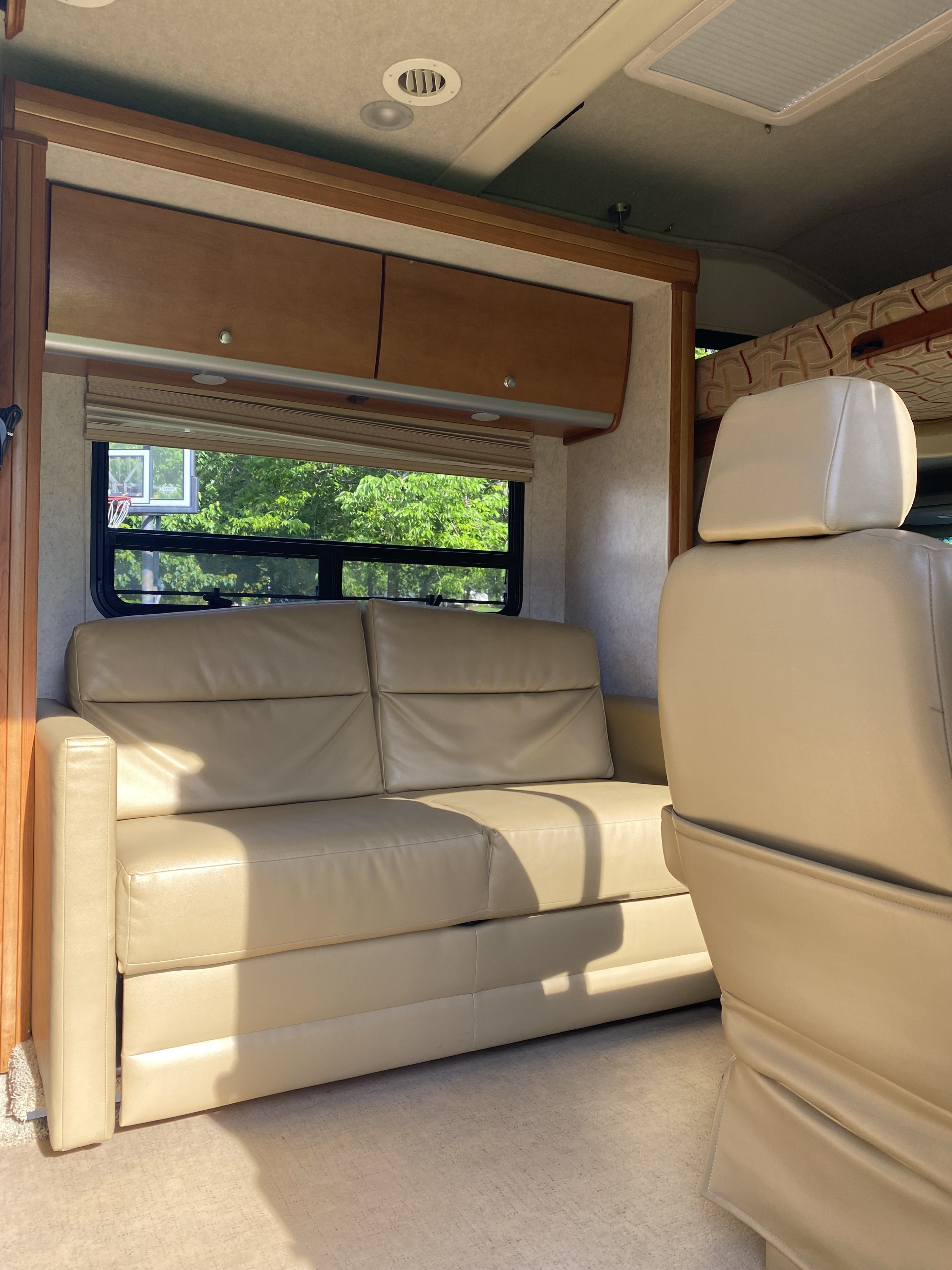 An RV rental interior showing a tan sofa, wood cabinets, and a window view revealing green trees and a basketball hoop outside.. Winnebago View 2010