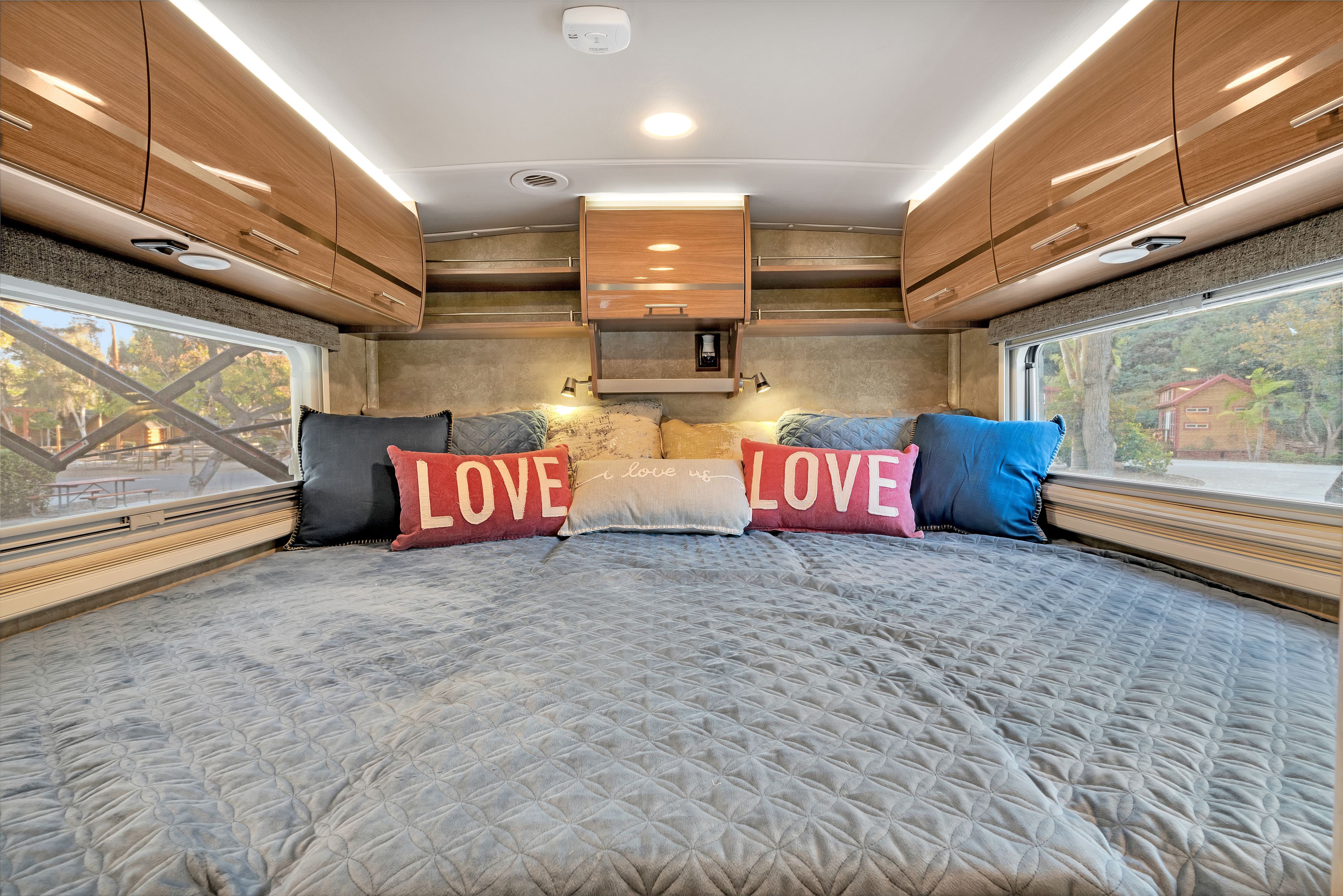 Comfy RV rental bedroom with a large bed, "LOVE" pillows, overhead cabinets, and scenic windows on both sides.. Winnebago Via 2015