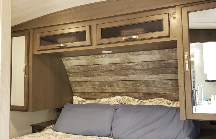 Cozy RV rental bedroom showcasing a rustic headboard, overhead cabinets with modern hardware, and a comfortable bed setup.. Keystone RV Cougar 2021