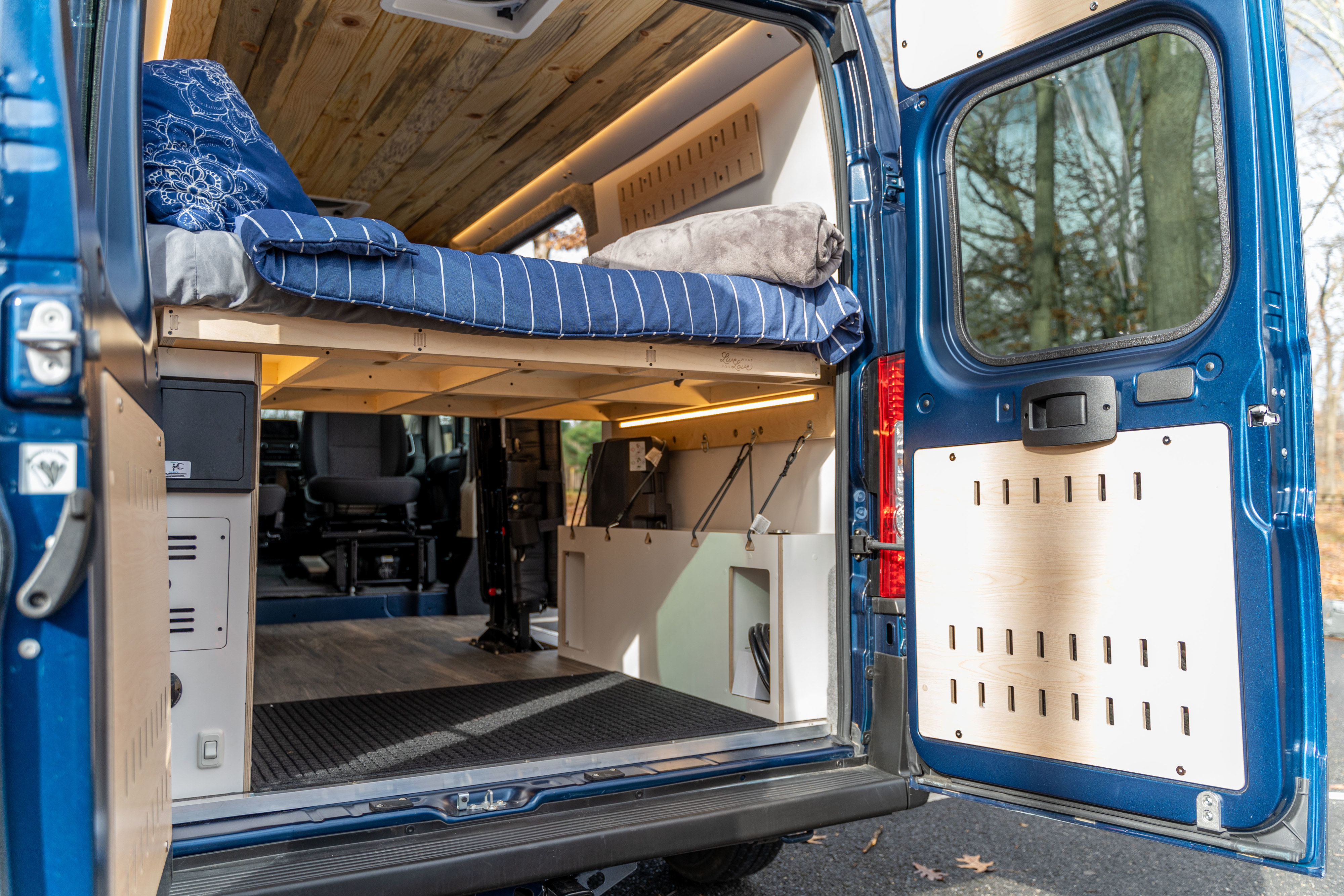 Inside a blue RV rental with open rear doors. Features a raised bed with blue bedding, custom wood ceiling, and lower storage.. Other Other 2022