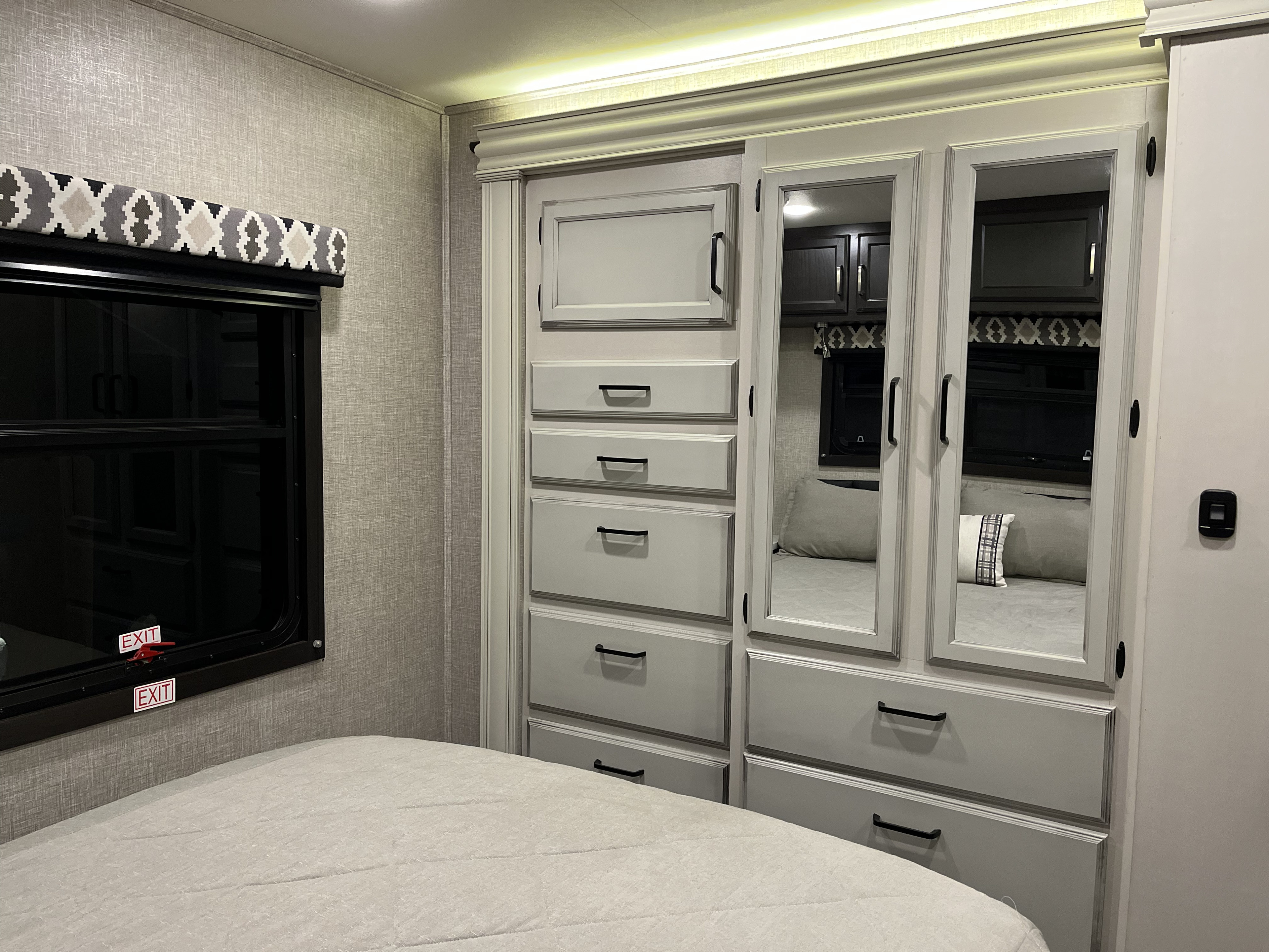 Bright RV rental bedroom: bed, window with patterned valance, and gray mirrored wardrobe with overhead lighting.. Jayco Redhawk 2022