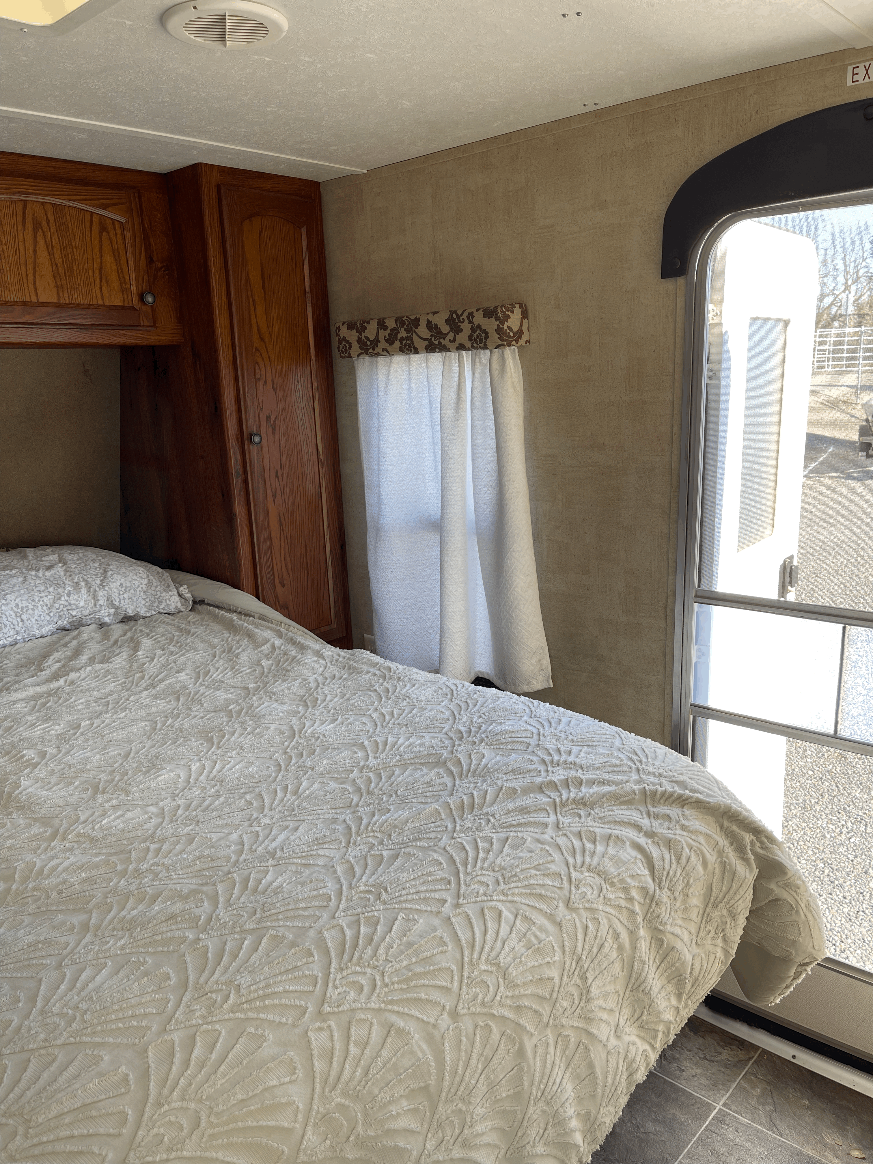 An RV rental bedroom with a white bed, wood cabinets, a patterned curtain, and an open door leading outside.. Keystone RV Hideout 2013