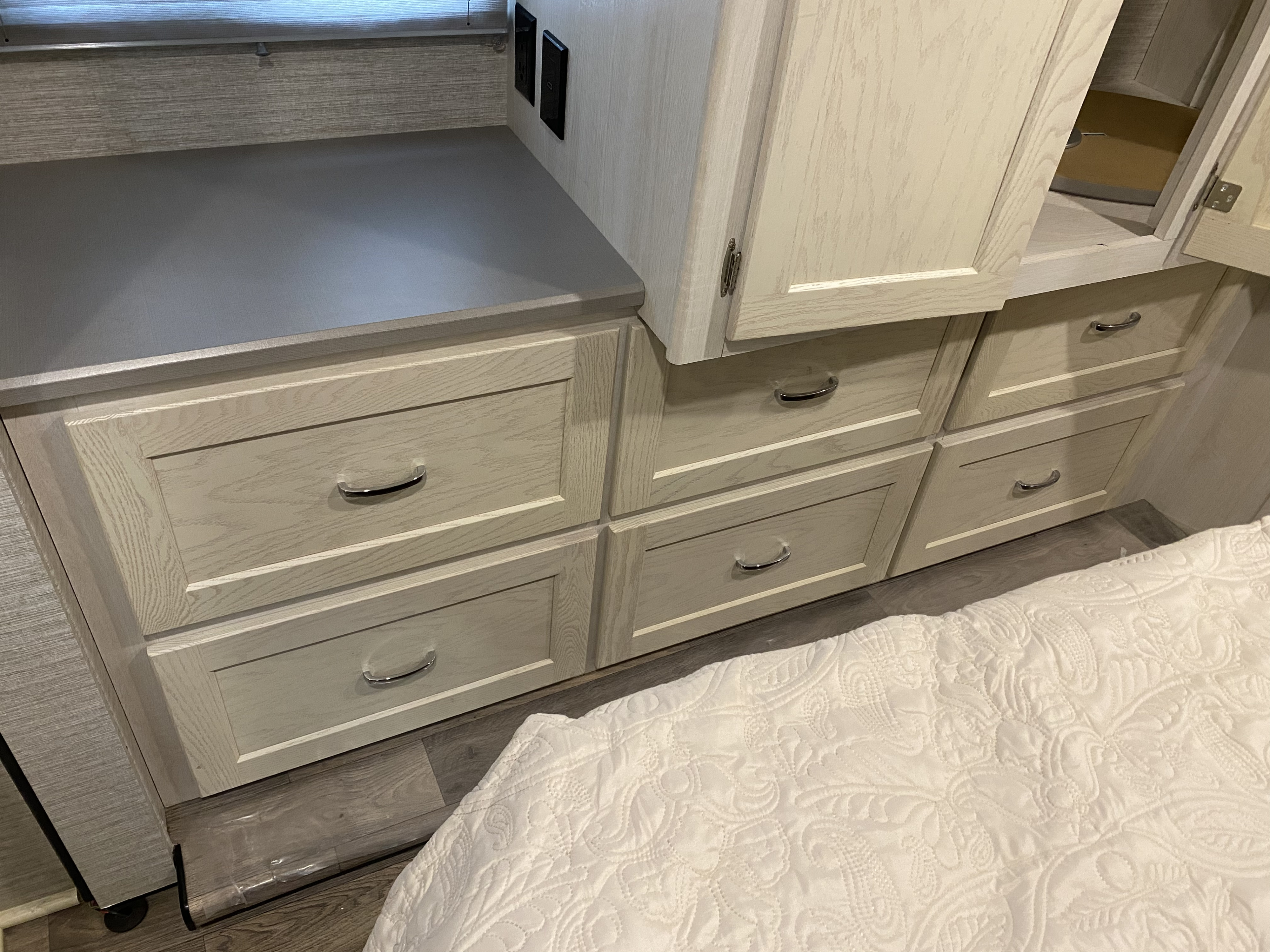 RV rental bedroom storage: Light wood-grain dresser with grey top, six drawers, and overhead cabinet. White quilted comforter visible.. Winnebago Intent 2020