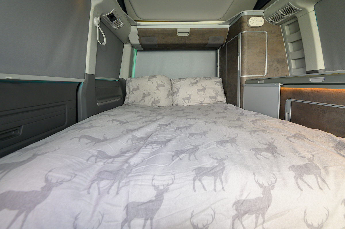 Inside a comfortable RV rental: A made-up bed with deer print bedding, built-in storage, and fridge.. Volkswagen California Ocean 2024