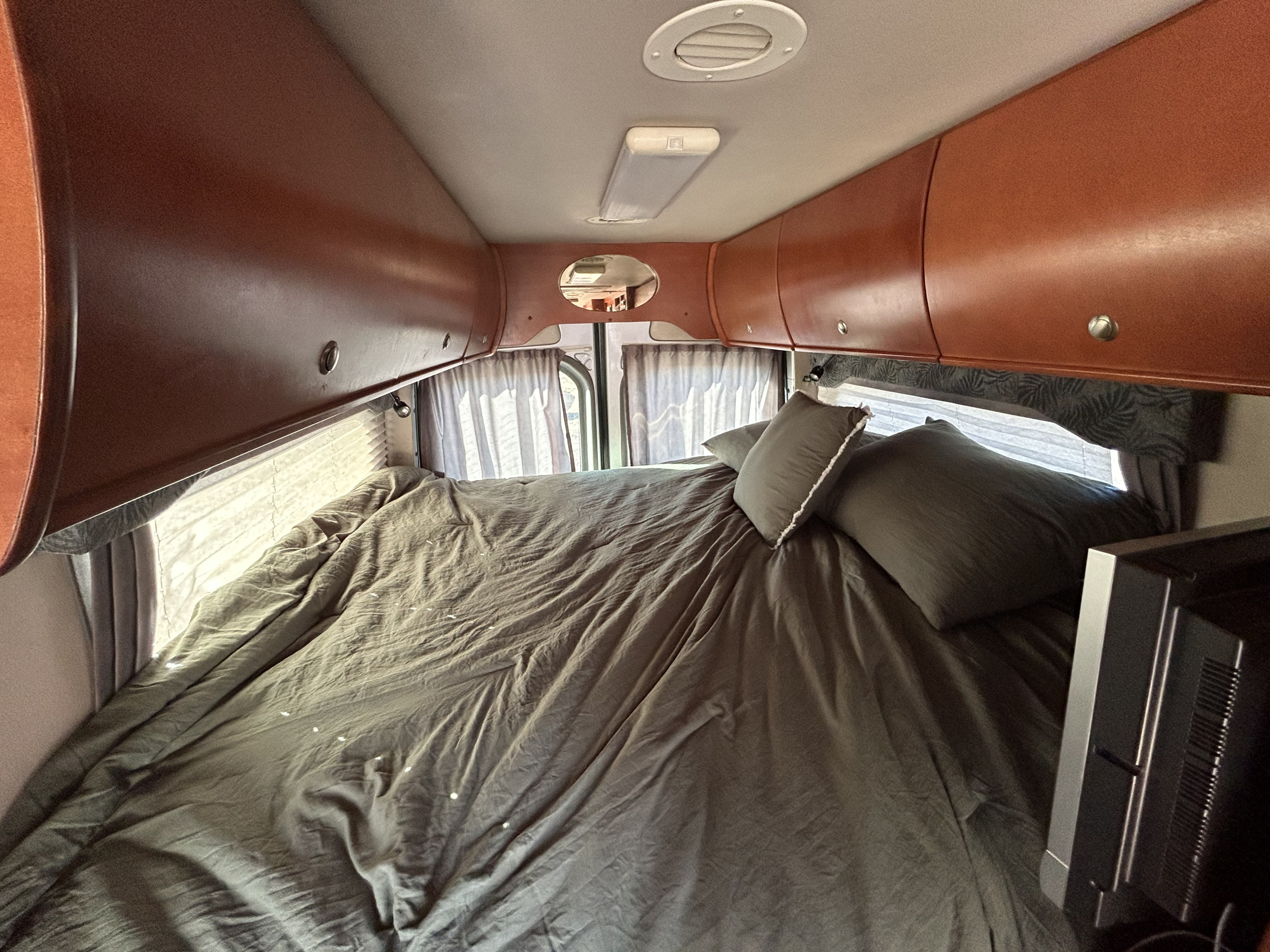 Cozy RV rental bedroom featuring olive green bedding, overhead storage, windows with curtains, and a small TV.. Mercedes-Benz Sprinter RV Motorhome Campervan 2008
