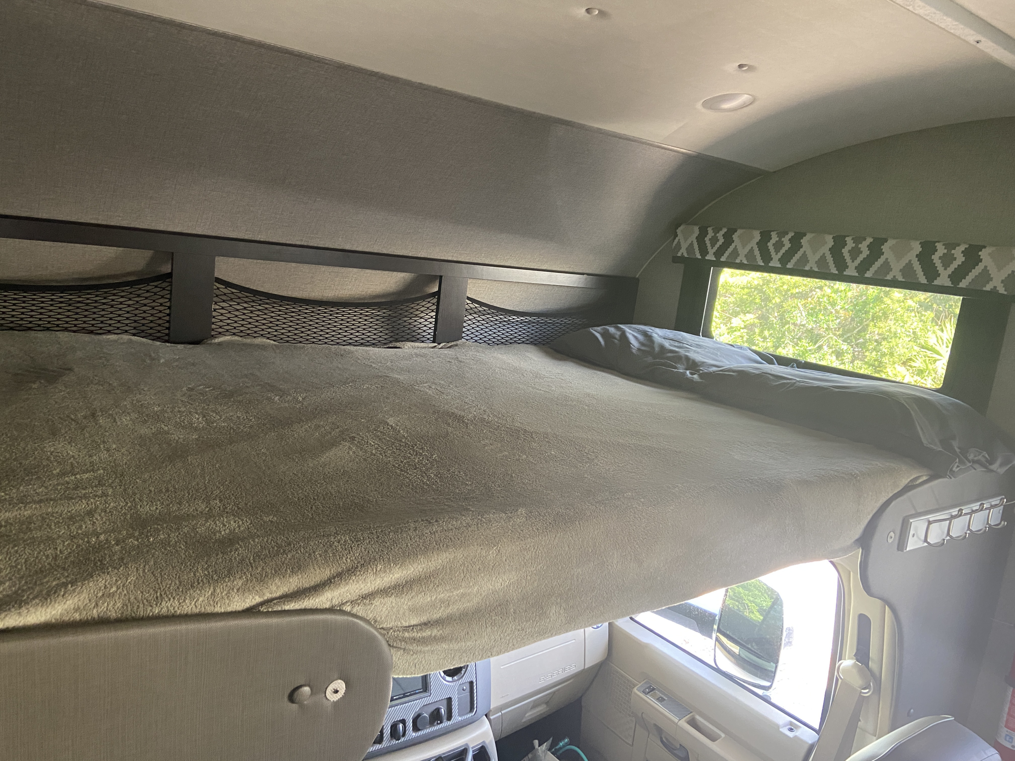 Cozy over-cab sleeping area in an RV rental, featuring a comfortable bed, mesh storage, and a scenic window view.. Jayco Greyhawk 2020