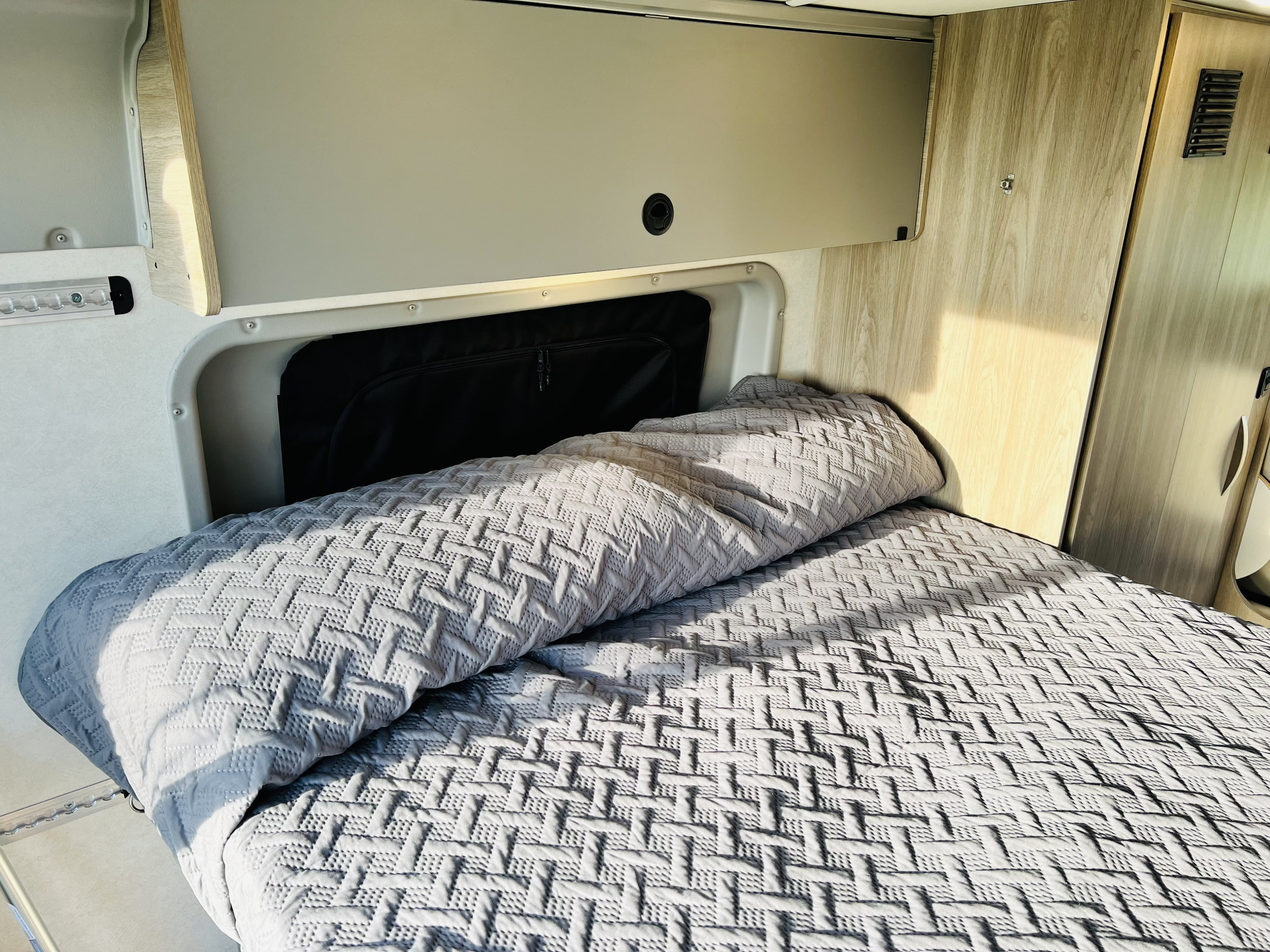 Cozy sleeping area in an RV rental, featuring a comfortable bed with textured grey bedding and overhead storage.. Winnebago Solis 2023