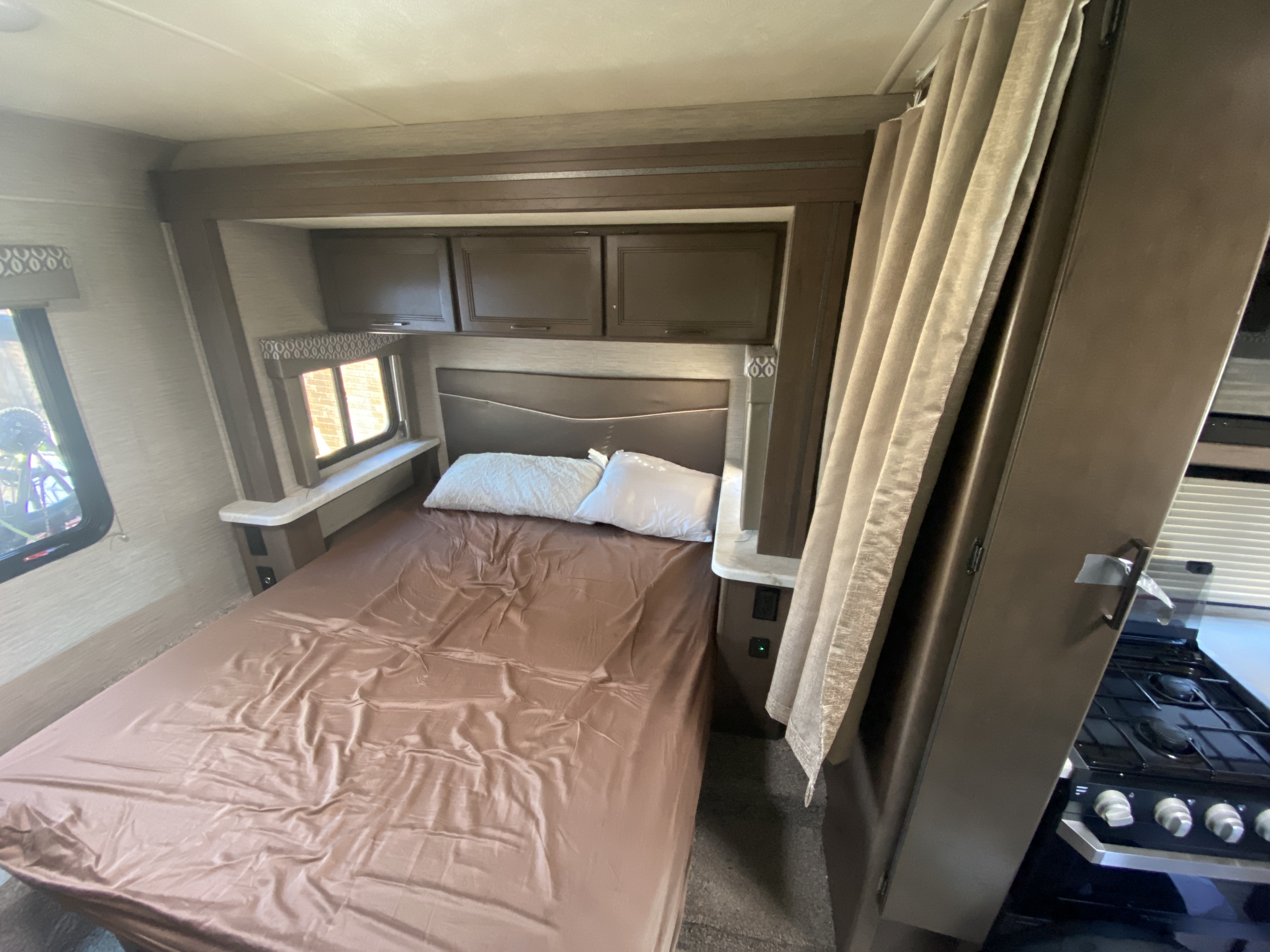 RV rental bedroom: A comfortable bed with brown satin sheets, overhead storage cabinets, window, and kitchen area nearby.. Thor Freedom Elite 2021