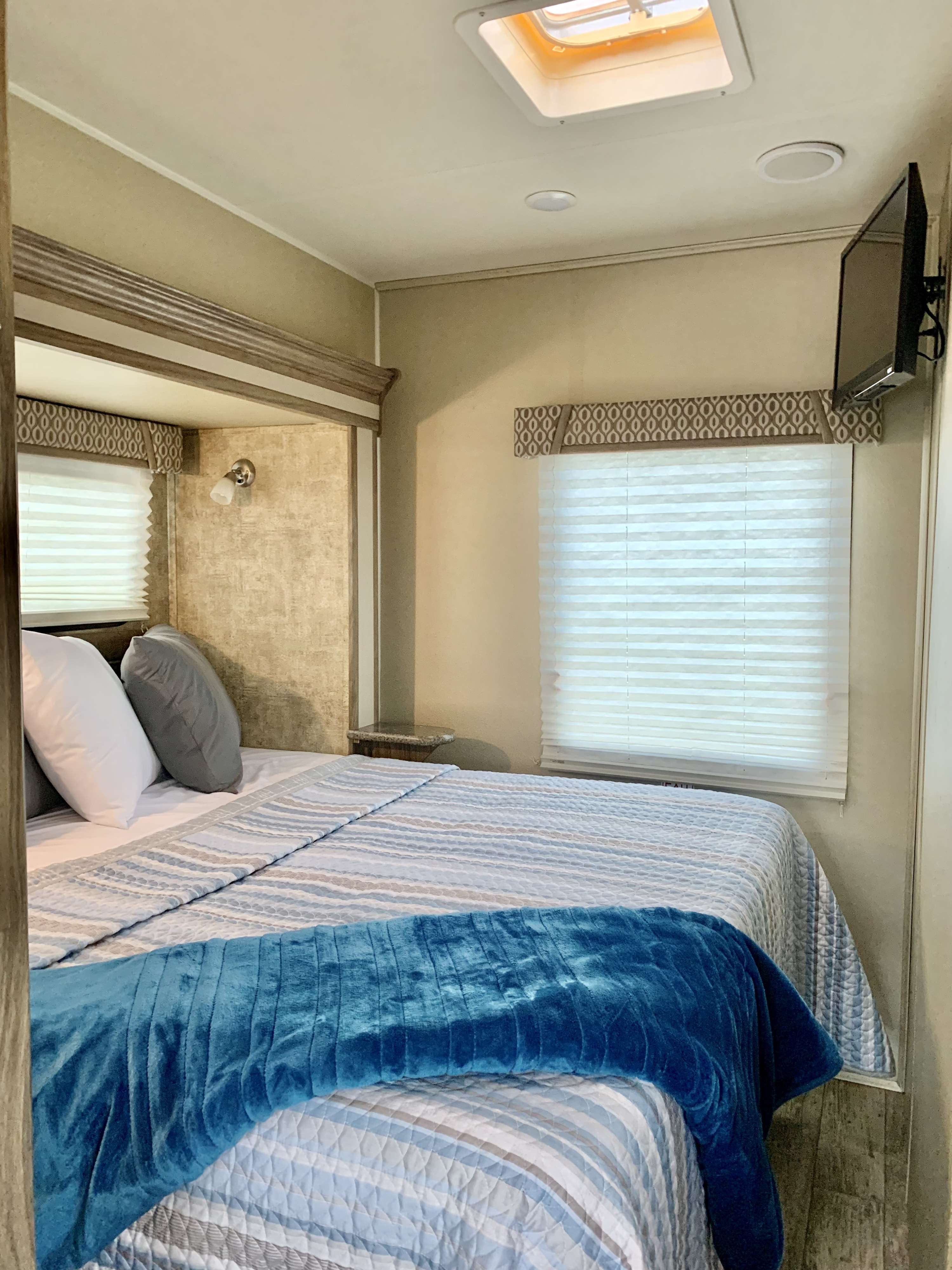 Comfortable RV rental bedroom with blue/grey bedding, window blinds, and TV. Features overhead lighting and a skylight for bright travel.. Jayco Envoy 100 2018