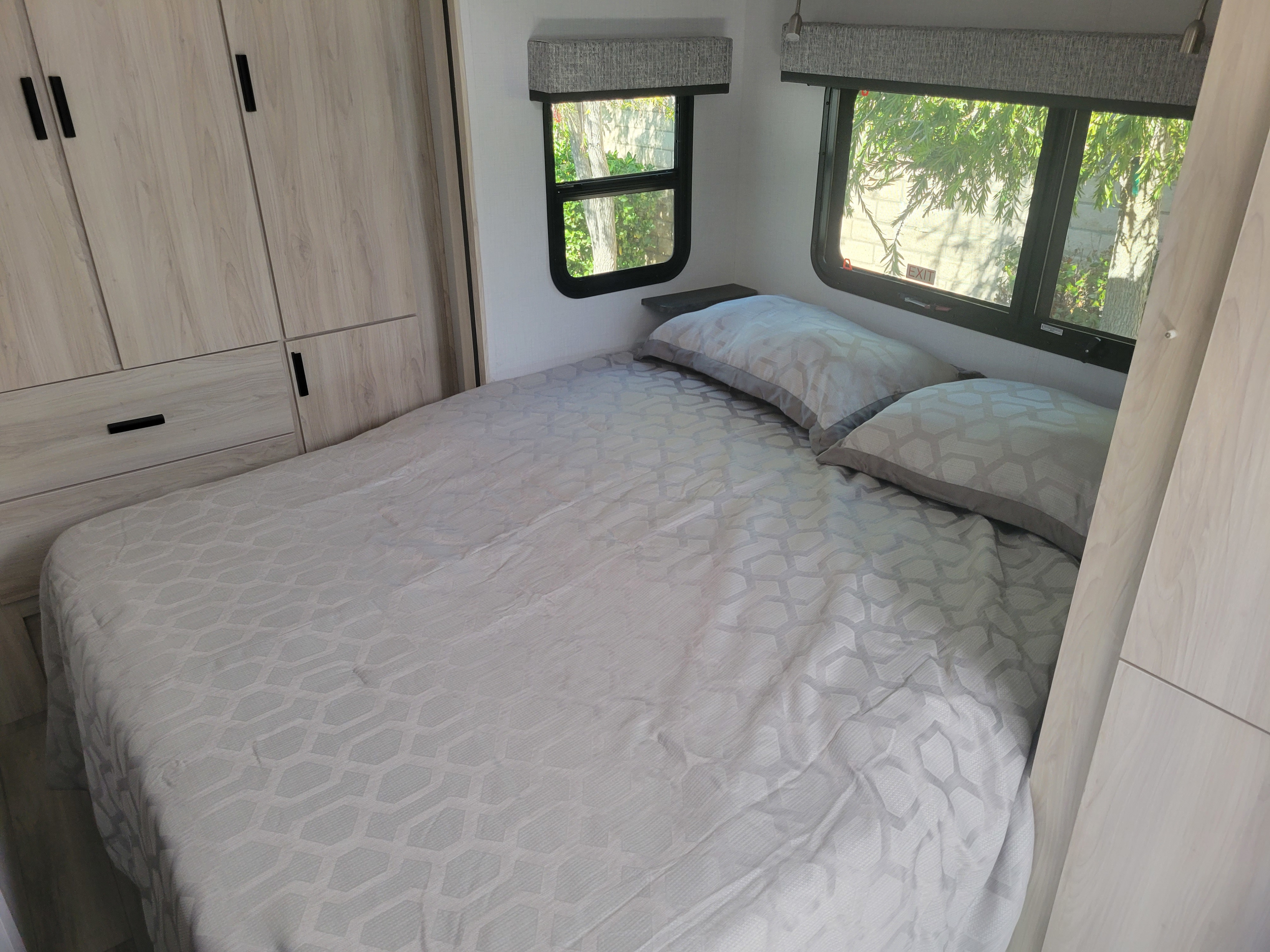 Cozy rv rental bedroom with a bed, light wood cabinets, and two windows looking out to green trees. Perfect for your travels.. Forest River Forester 2023