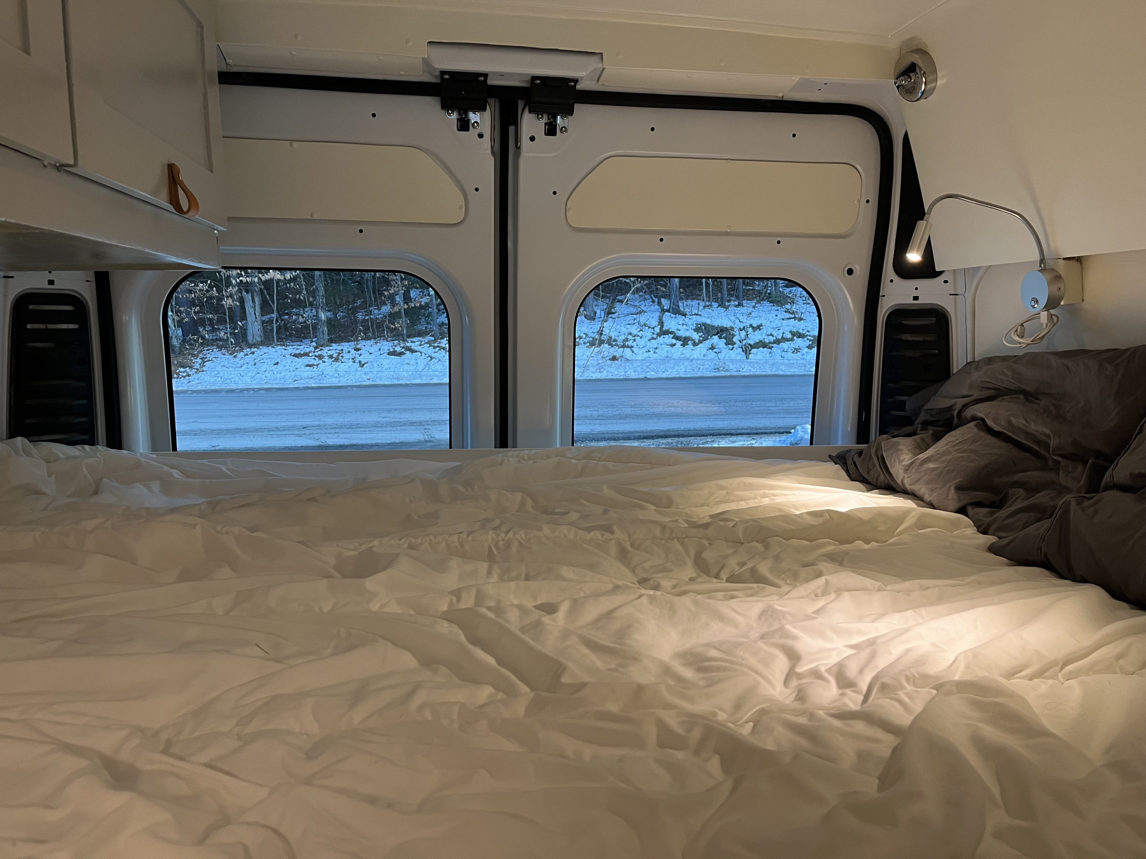 Cozy rv rental interior: A bed with white sheets and gray bedding, offering a view of a snowy road and forest for winter travel.. Ram Promaster 2022