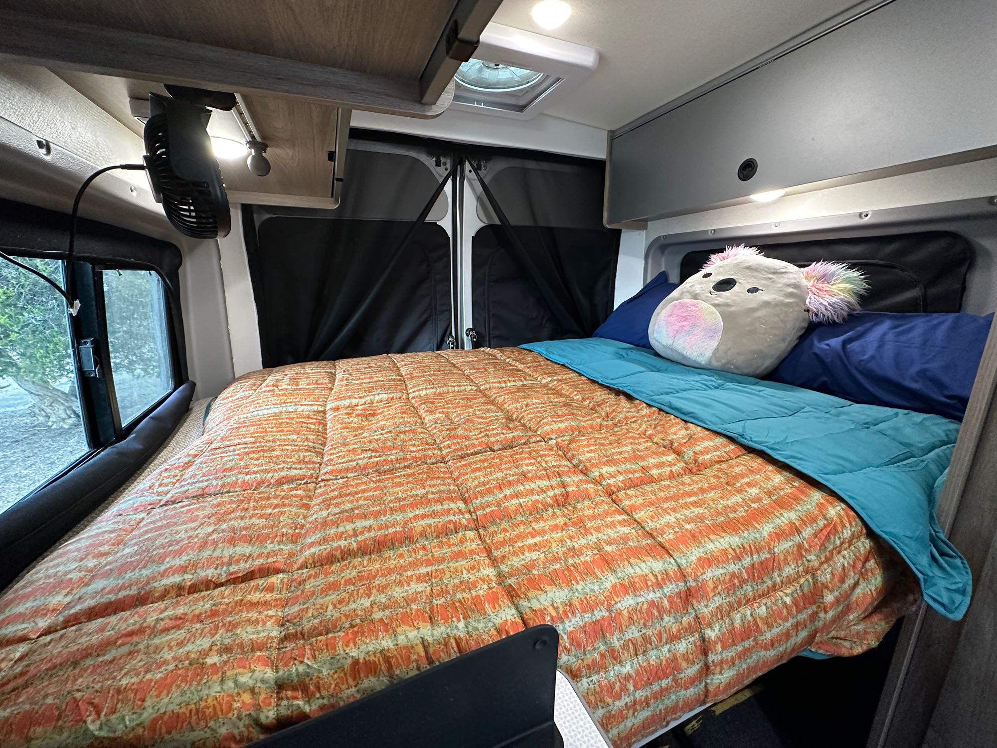 Comfortable RV rental bedroom: cozy bed with colorful bedding, fan, overhead storage, and privacy window covers.. Winnebago Solis 2022