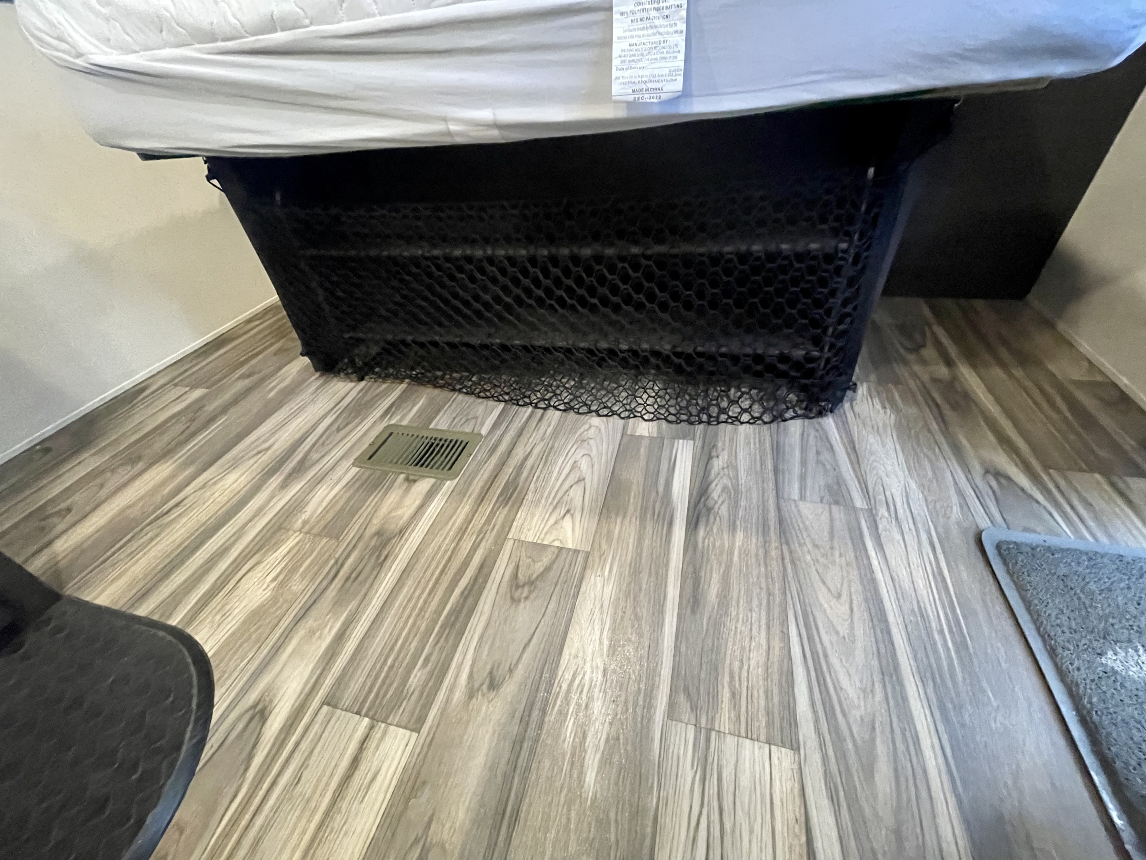 RV rental bed with under-bed mesh storage net, visible mattress label, and floor vent on wood-look flooring.. Forest River EVO T2250 2019