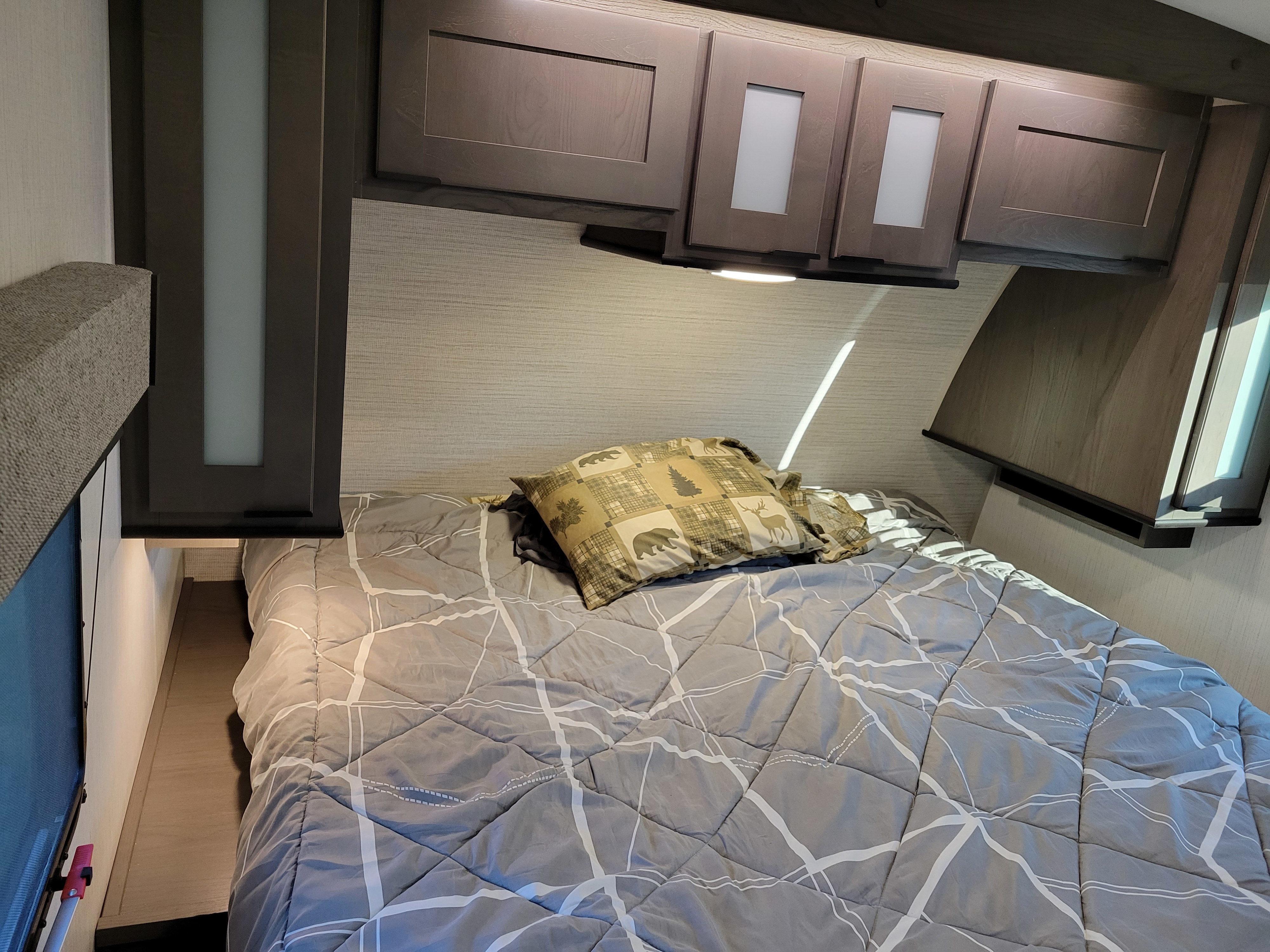 Inside an RV rental: a neat bedroom with a patterned gray comforter, rustic-themed pillow, and overhead wood cabinets.. Cruiser RV MPG Ultra Lite 2022