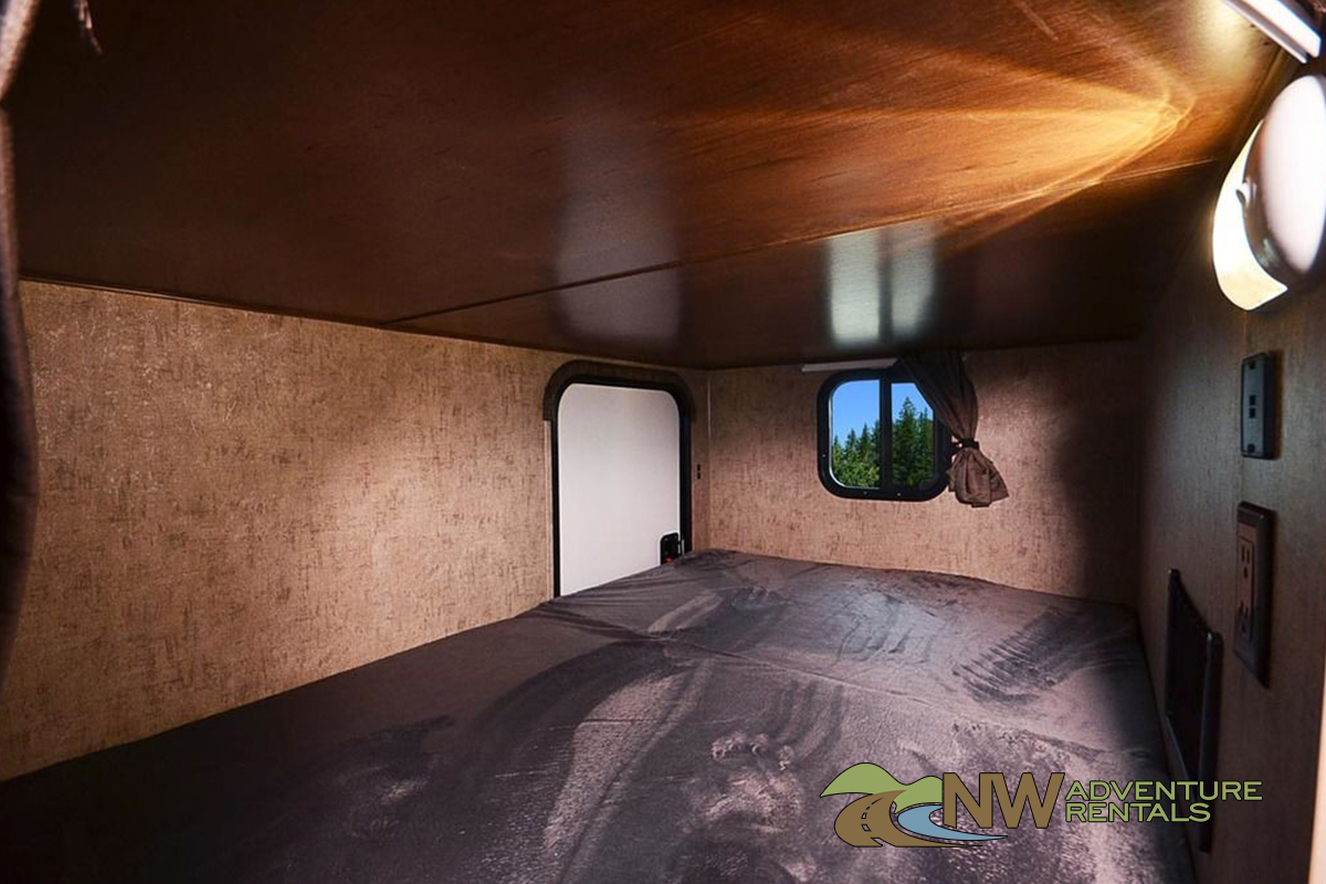 RV rental bedroom with a dark bed, beige textured walls, and a window framing lush trees and blue sky. Door on left.. Outdoors Rv Manufacturing Timber Ridge 23DBS Mountain 2023