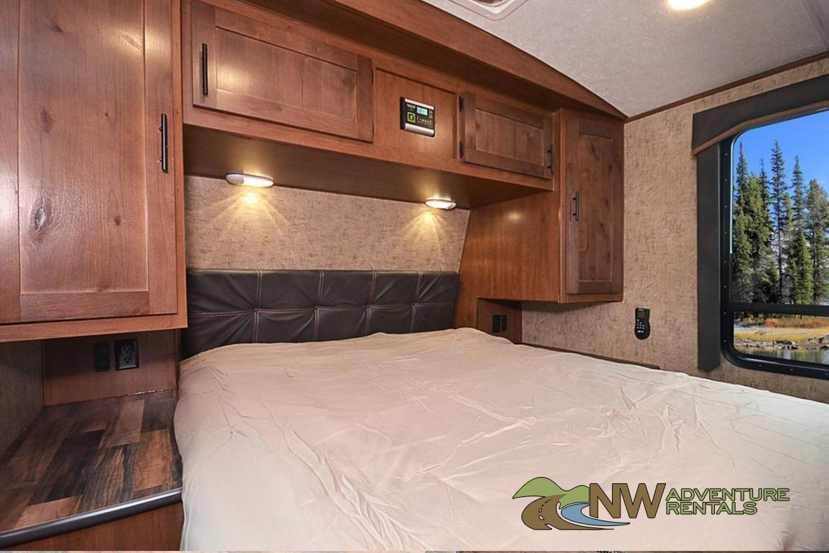 RV rental's cozy bedroom, showing overhead cabinets, reading lights, and a window with a scenic view of trees and water.. Outdoors Rv Manufacturing Timber Ridge 23DBS Mountain 2023
