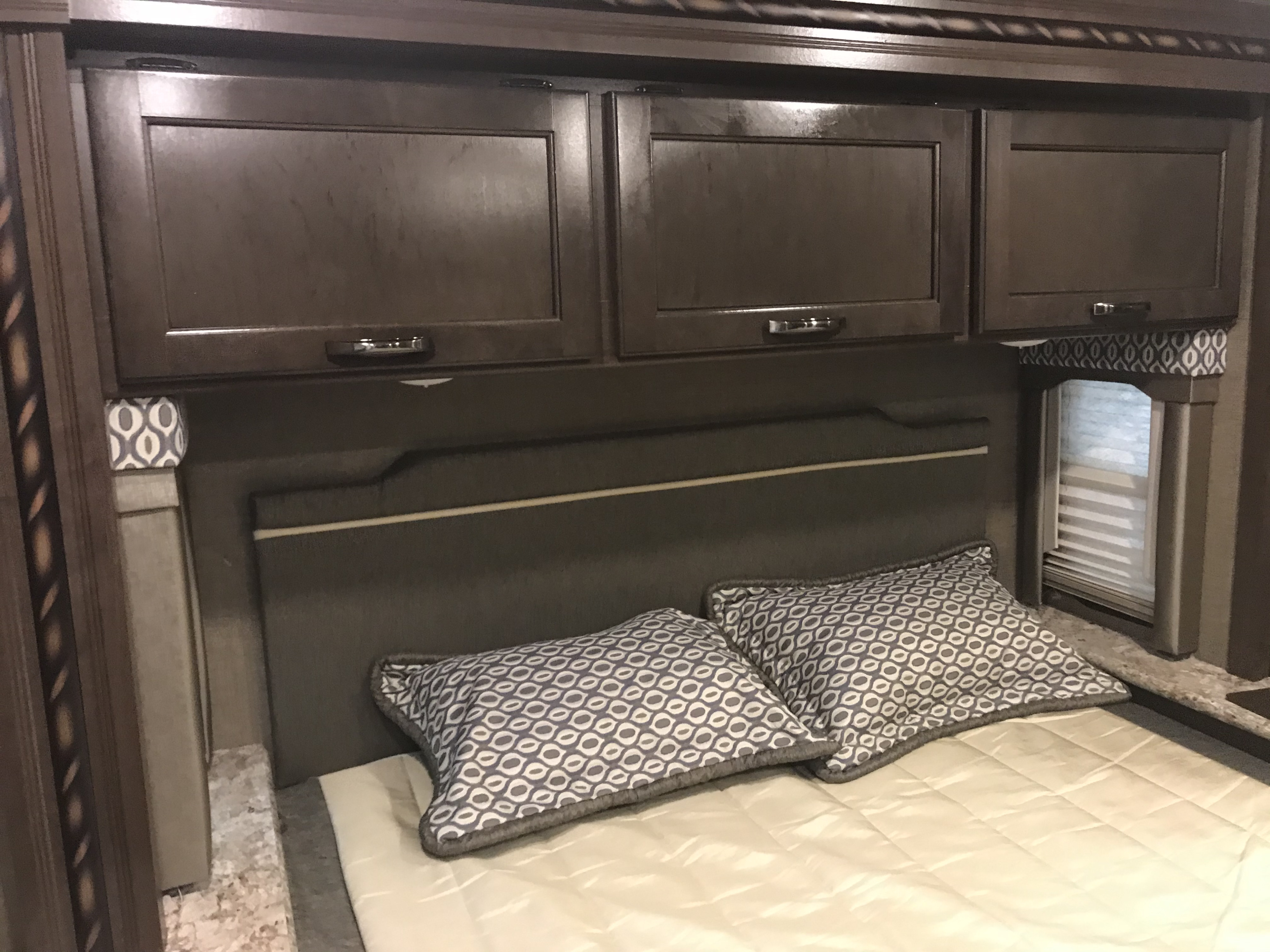 Cozy RV rental bedroom features dark wood overhead cabinets, a padded headboard, and a bed with patterned pillows.. Thor Freedom Elite 2018