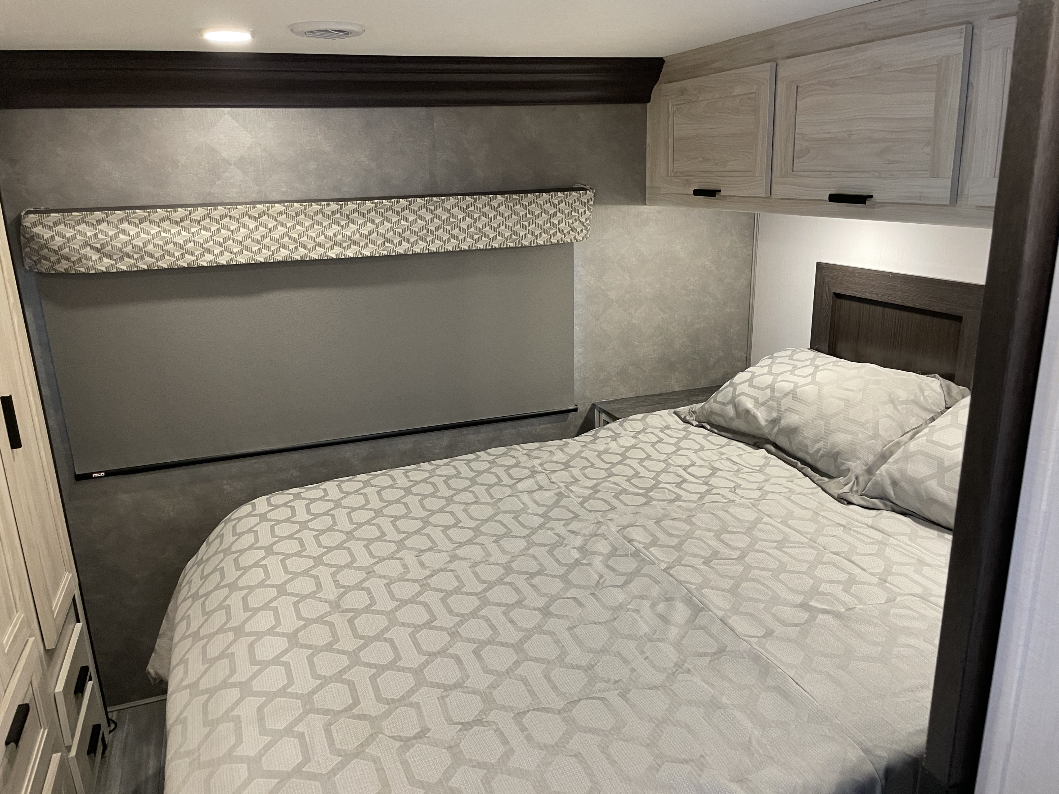 This modern RV rental bedroom offers a comfortable bed with grey patterned bedding, light wood cabinets, and a pull-down shade.. Forest River Forester 3251DSF 2023
