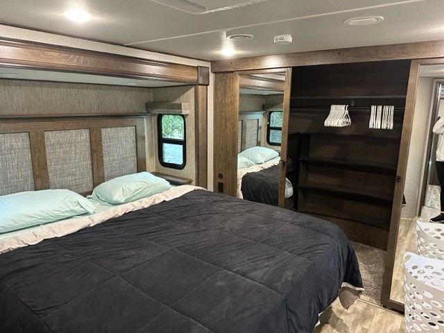 Interior of an RV rental bedroom showing a large bed with light blue pillows, wooden headboard, and open closet.. Grand Design Solitude 2020