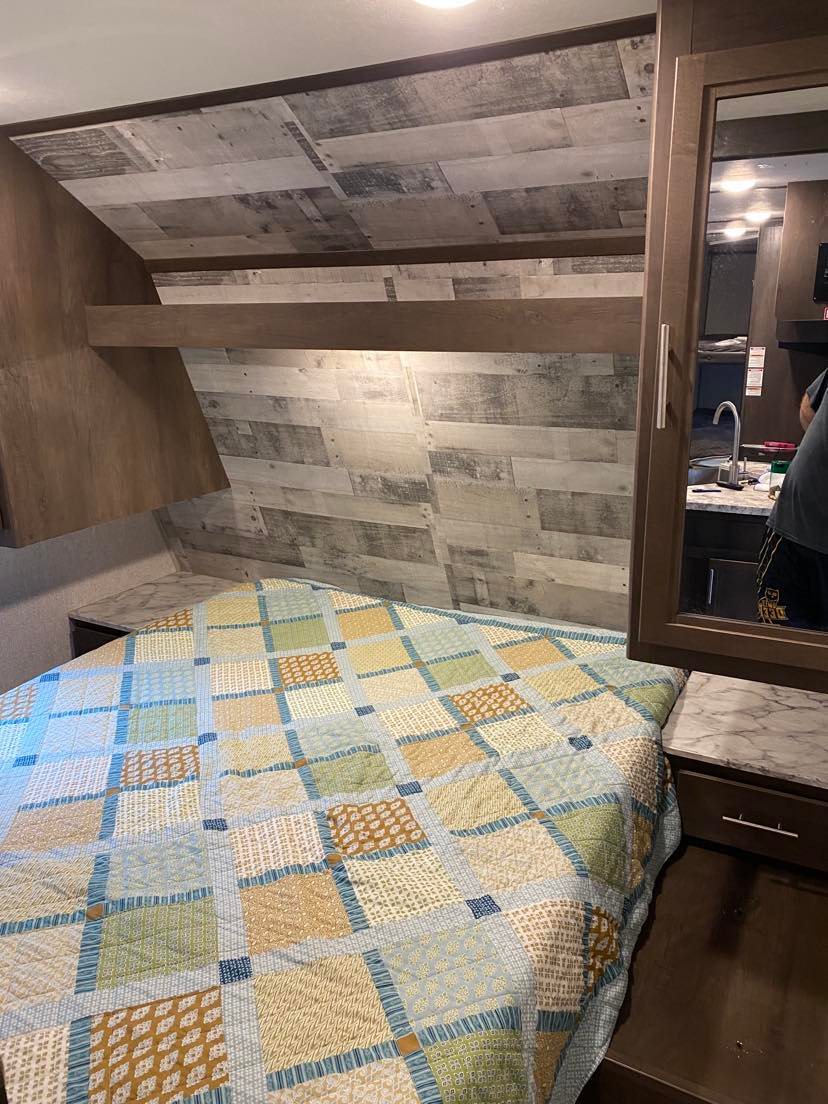 Cozy RV rental bedroom featuring a colorful patchwork quilt, wood-look walls, and a mirrored cabinet reflecting a kitchen area.. Dutchmen Coleman Lantern 2019