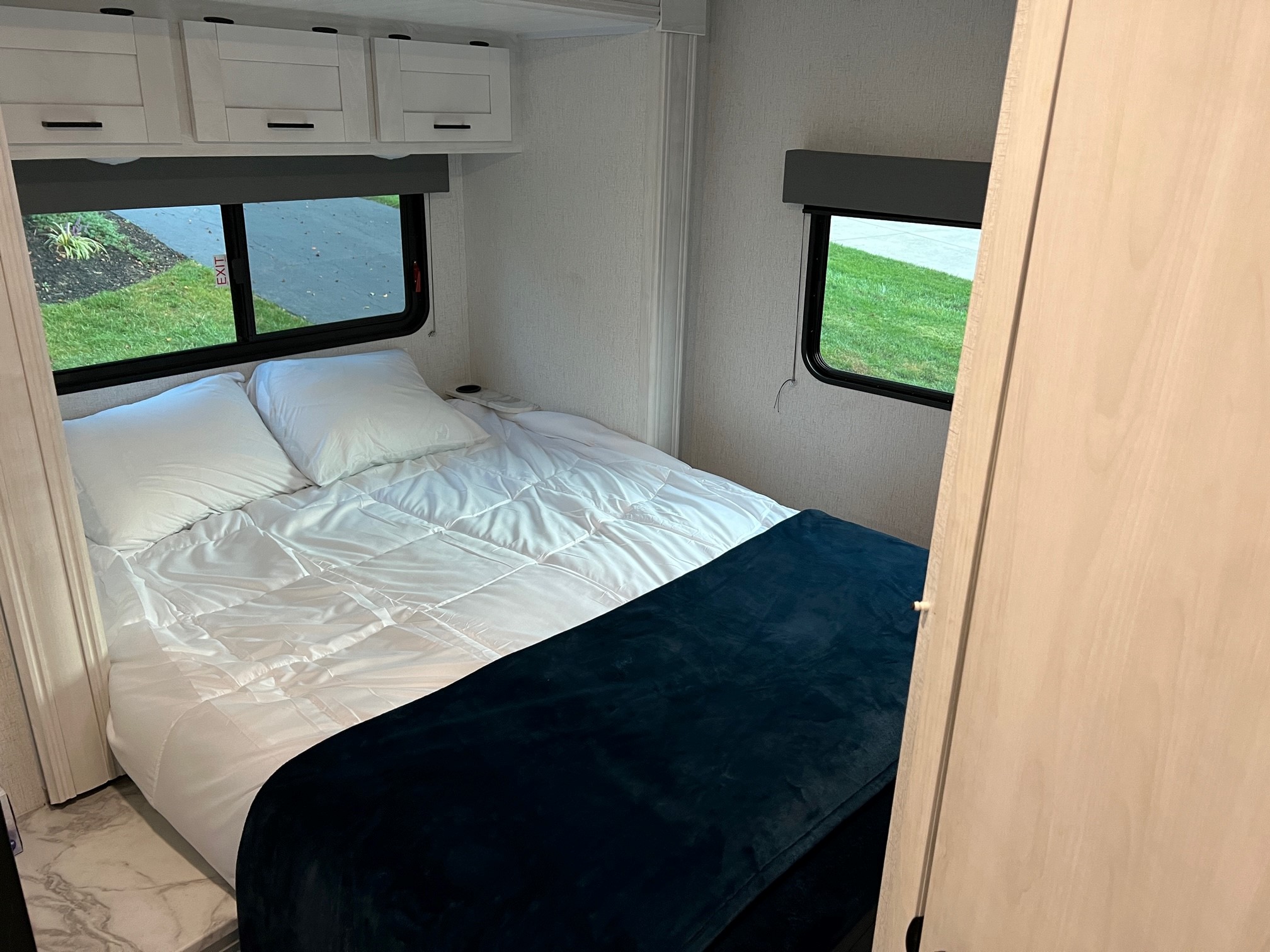 RV rental bedroom with a comfortable bed, white bedding, dark blue blanket, and two windows showing outdoor scenery.. East to West Entrada 2600DS 2022
