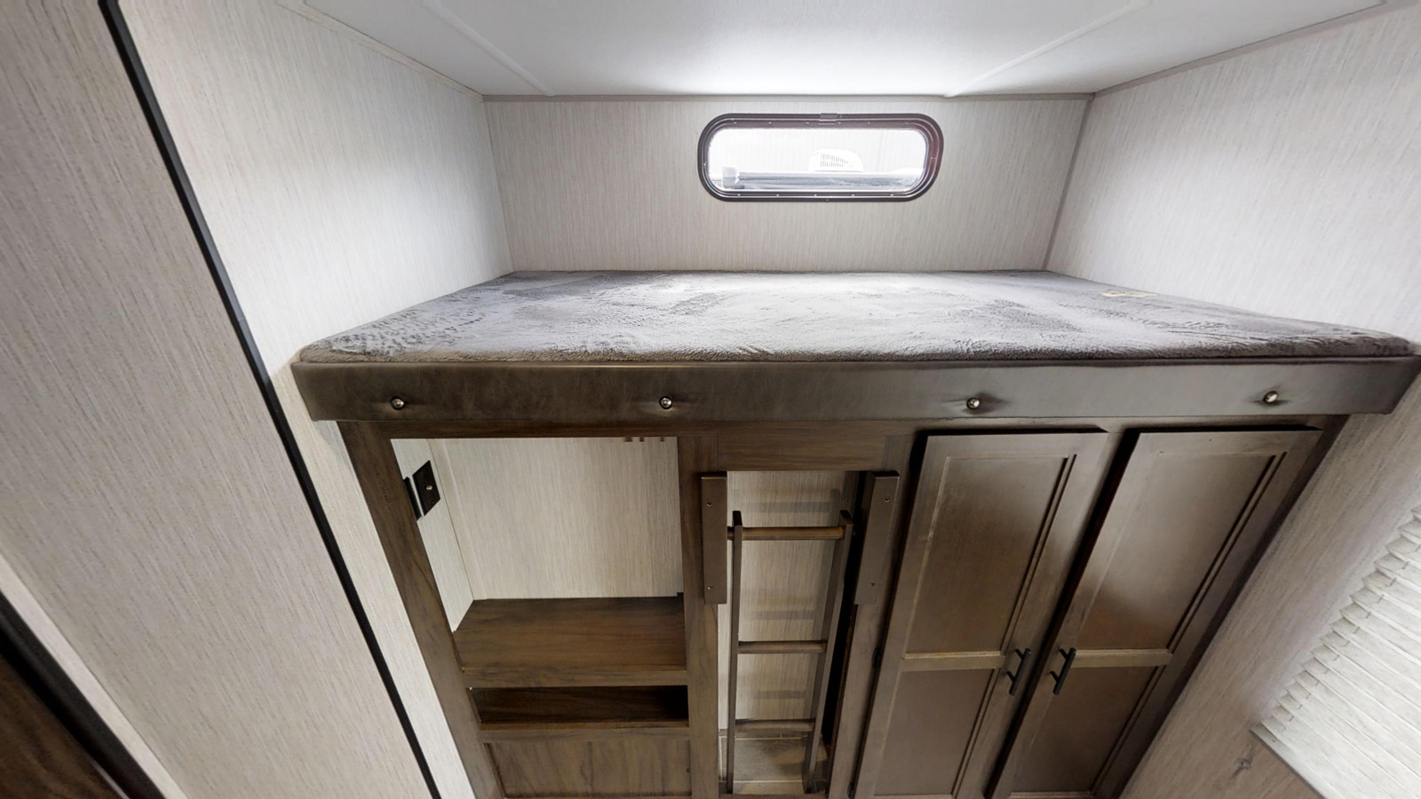 Inside an RV rental, a compact bunk bed sits above built-in storage shelves and cabinets, with a small oval window.. Forest River Cherokee 2019