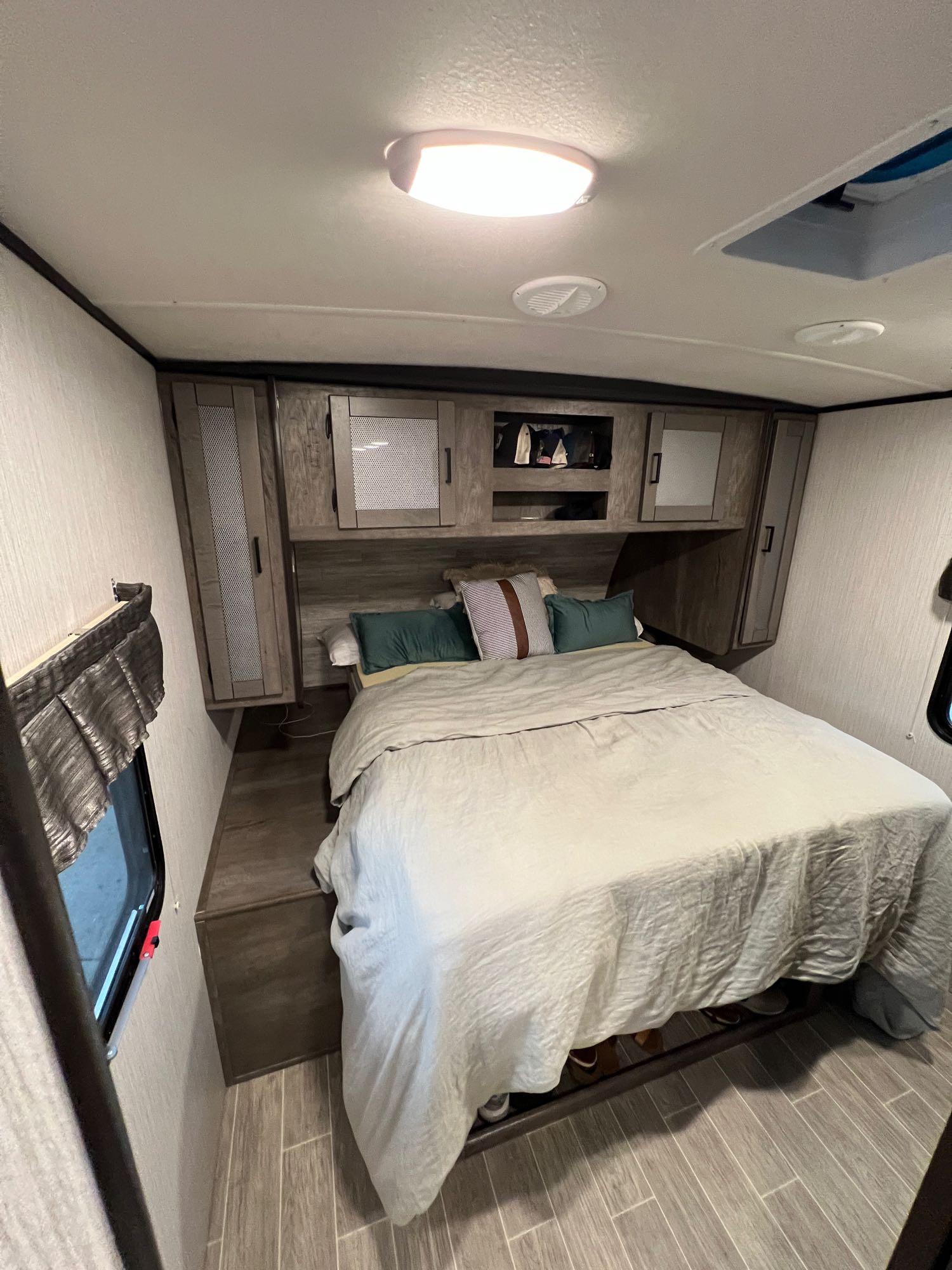 This RV rental bedroom features a large bed, overhead storage, and bright ceiling lights. Wood-look flooring and a window are visible.. Heartland Wilderness 2020